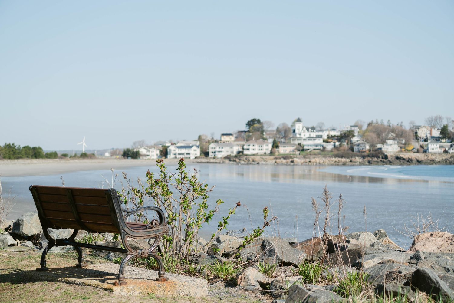 A wooden bench overlooks a calm river with a small town in the background on a clear day.