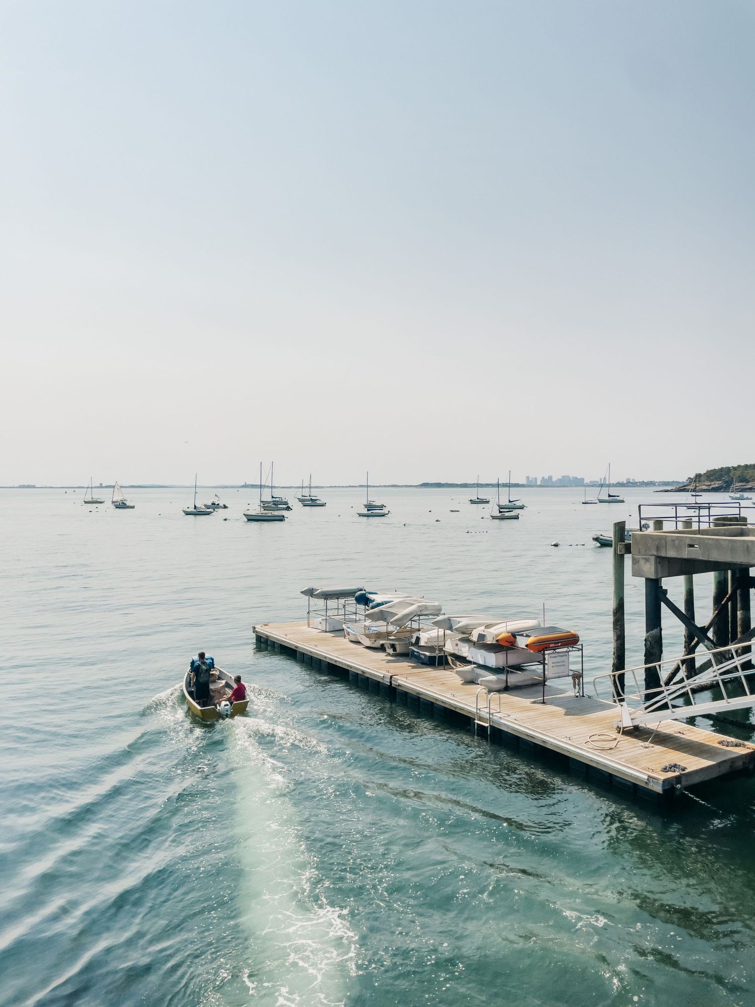 Small motorboat approaching wooden dock with sailboats anchored in calm harbor waters beyond.