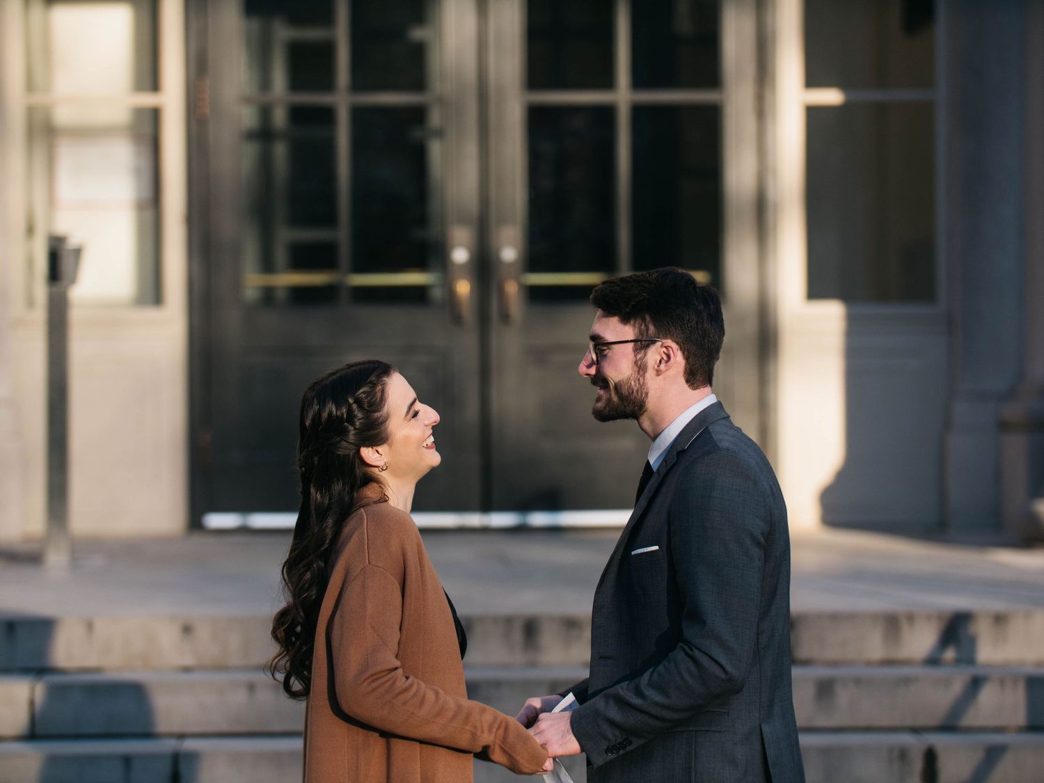 Couple holding hands and gazing at each other outside a grand stone building.
