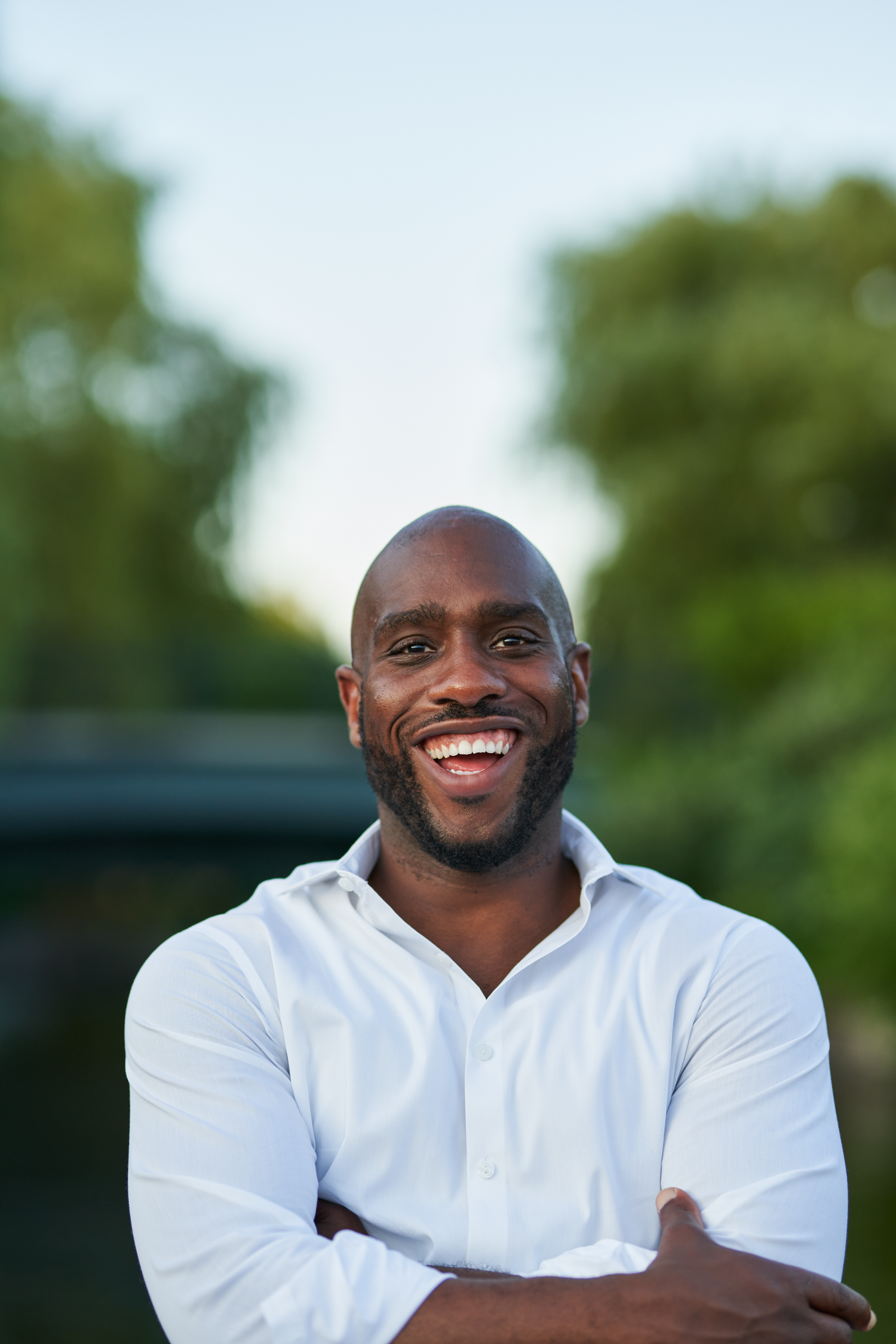 Smiling bald Black man with beard, arms crossed, wearing white shirt, standing outdoors near green trees and water.