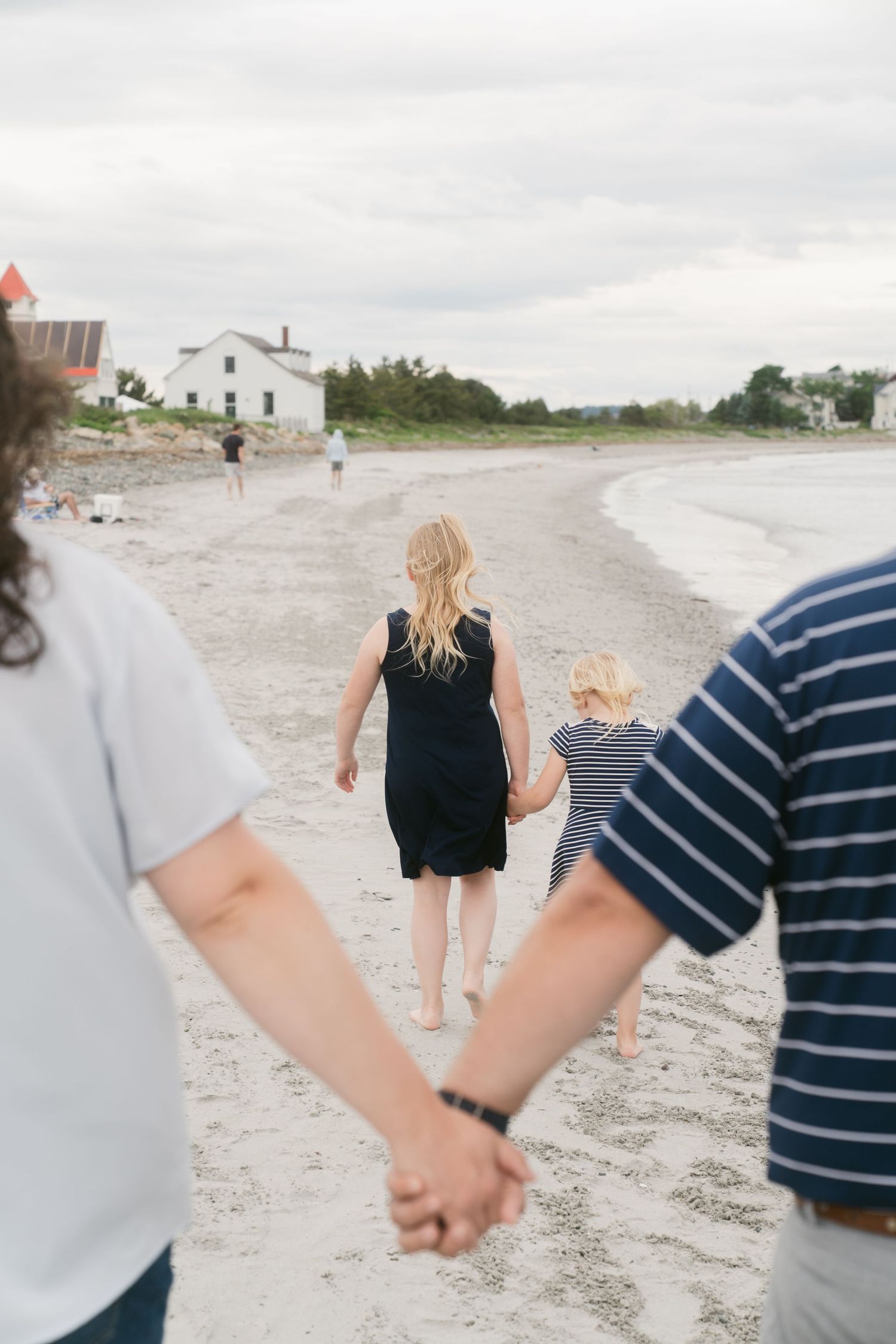 Family walking hand-in-hand on a sandy beach, with a woman in a black dress leading a child toward a white house.