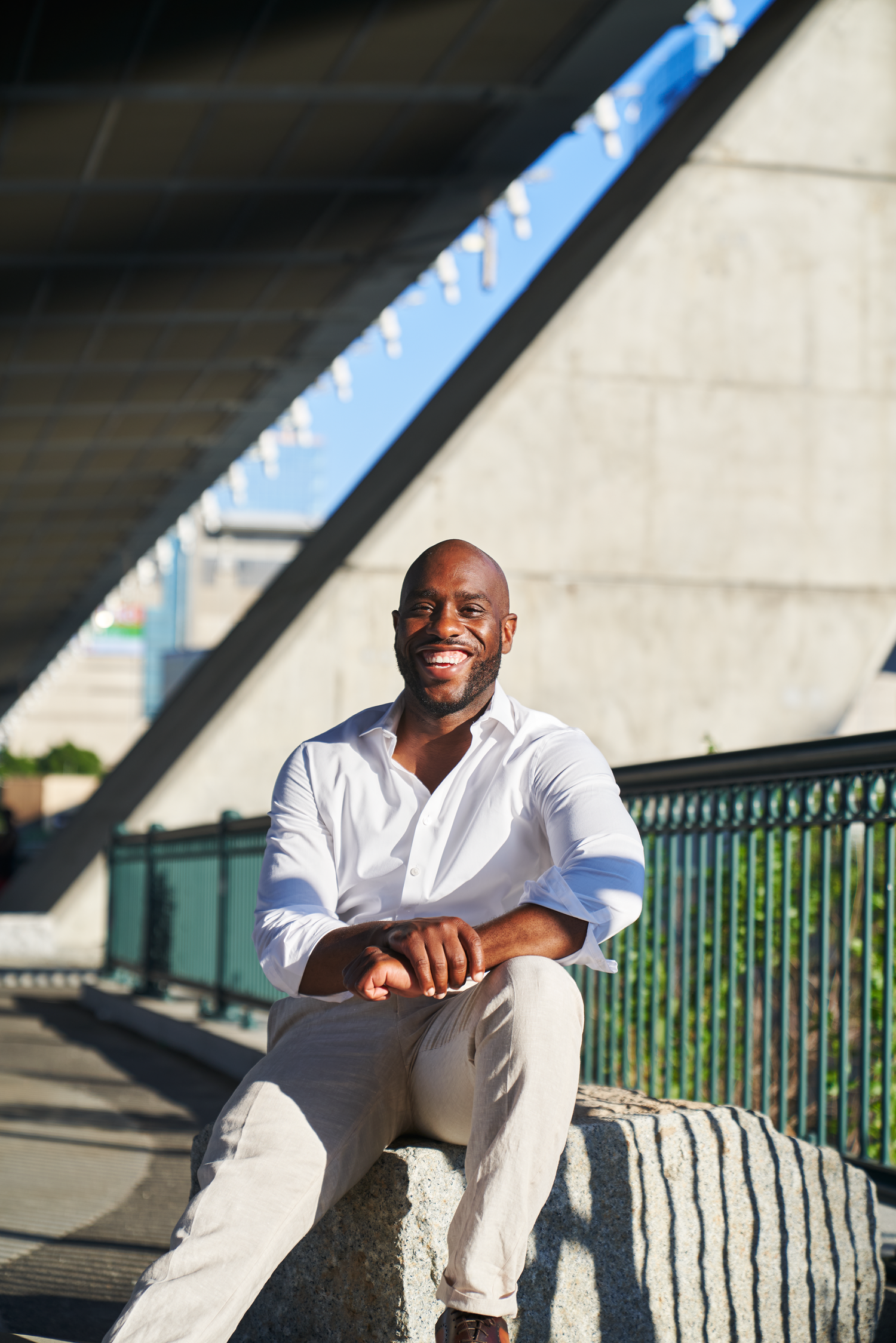 Smiling Black man in white shirt and khakis sitting outdoors on stone ledge under modern bridge structure.