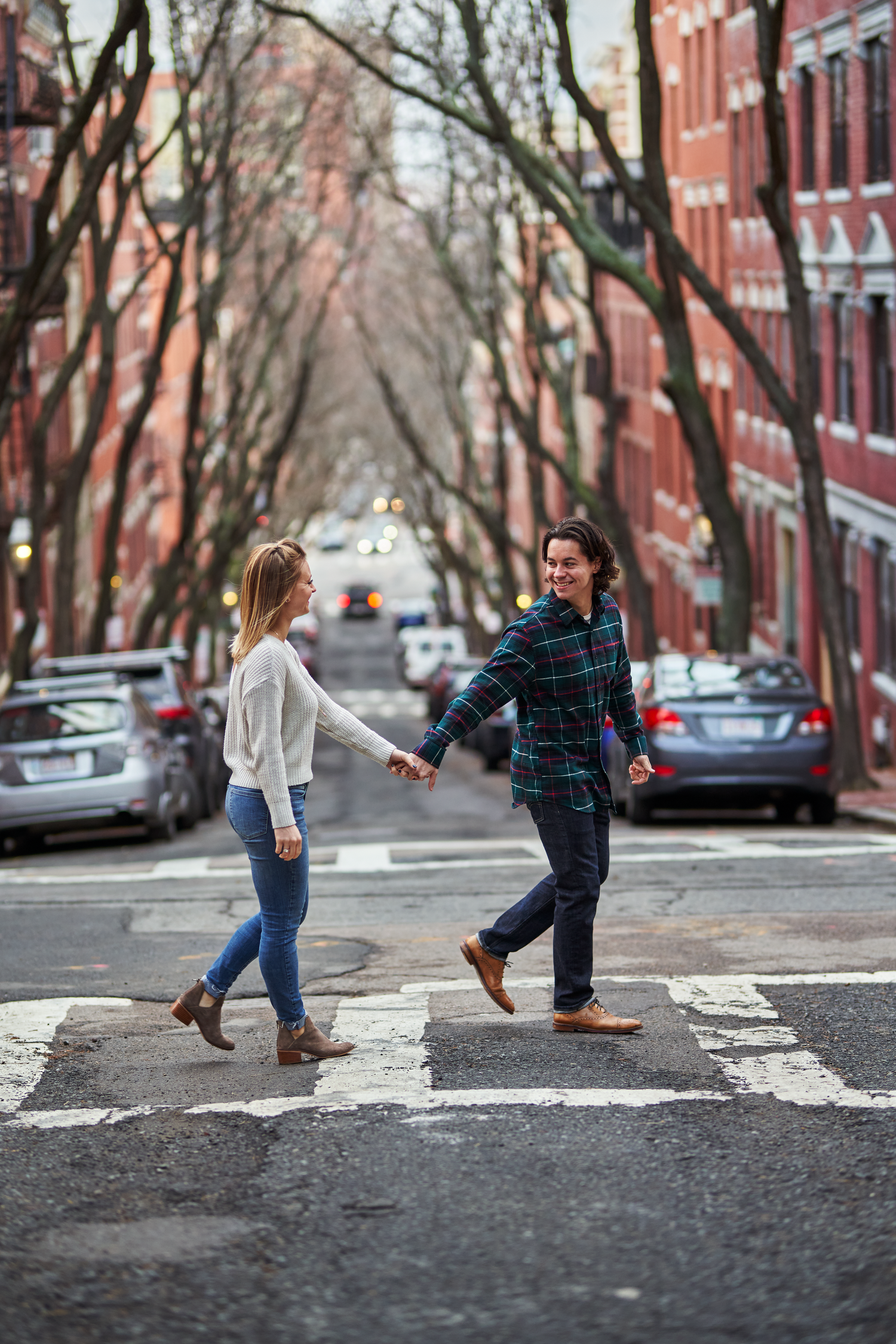 Joyful couple holding hands while crossing a tree-lined urban street with red brick buildings.