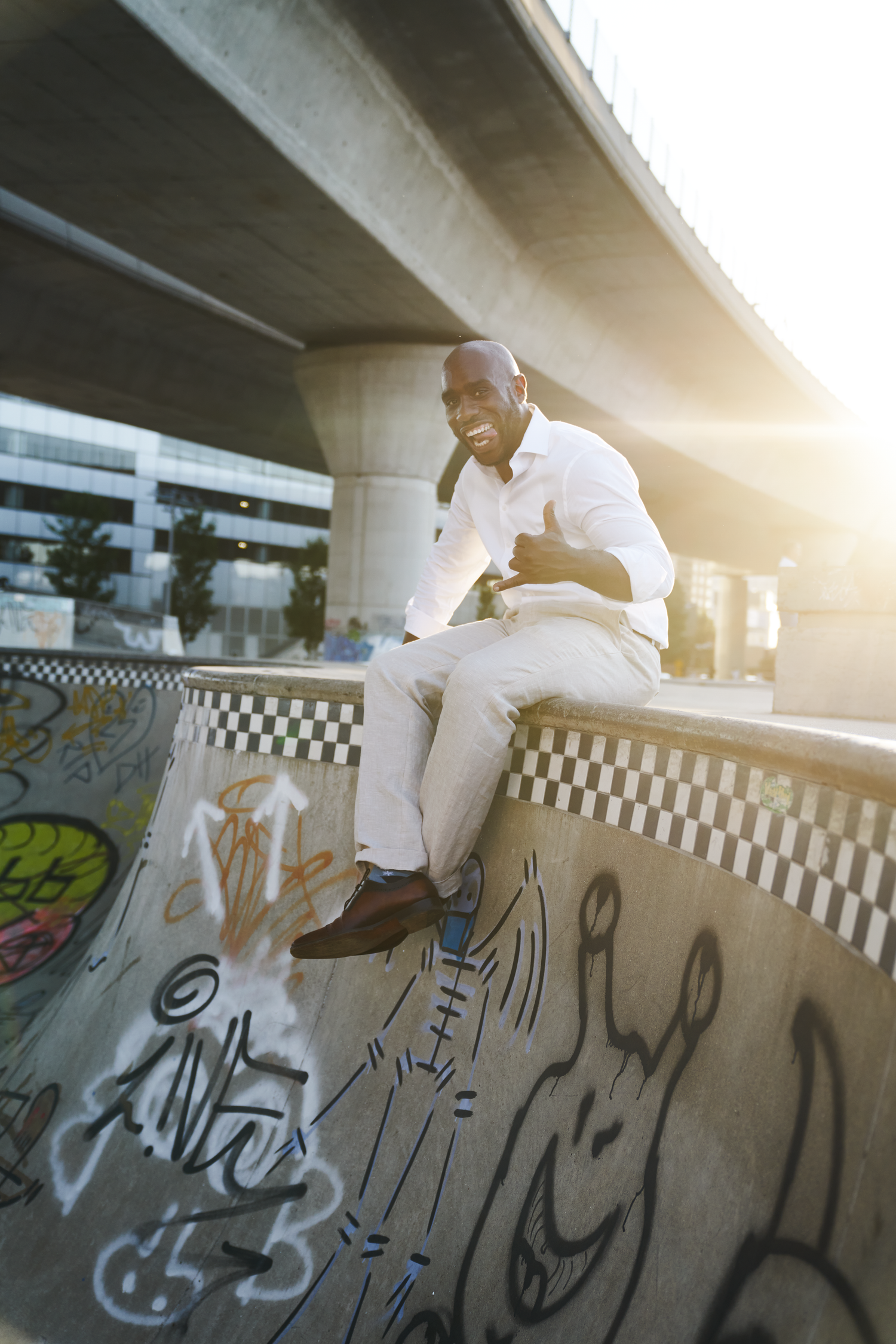 Smiling Black man in white shirt and khakis sitting on graffiti skate ramp under bridge in urban setting.
