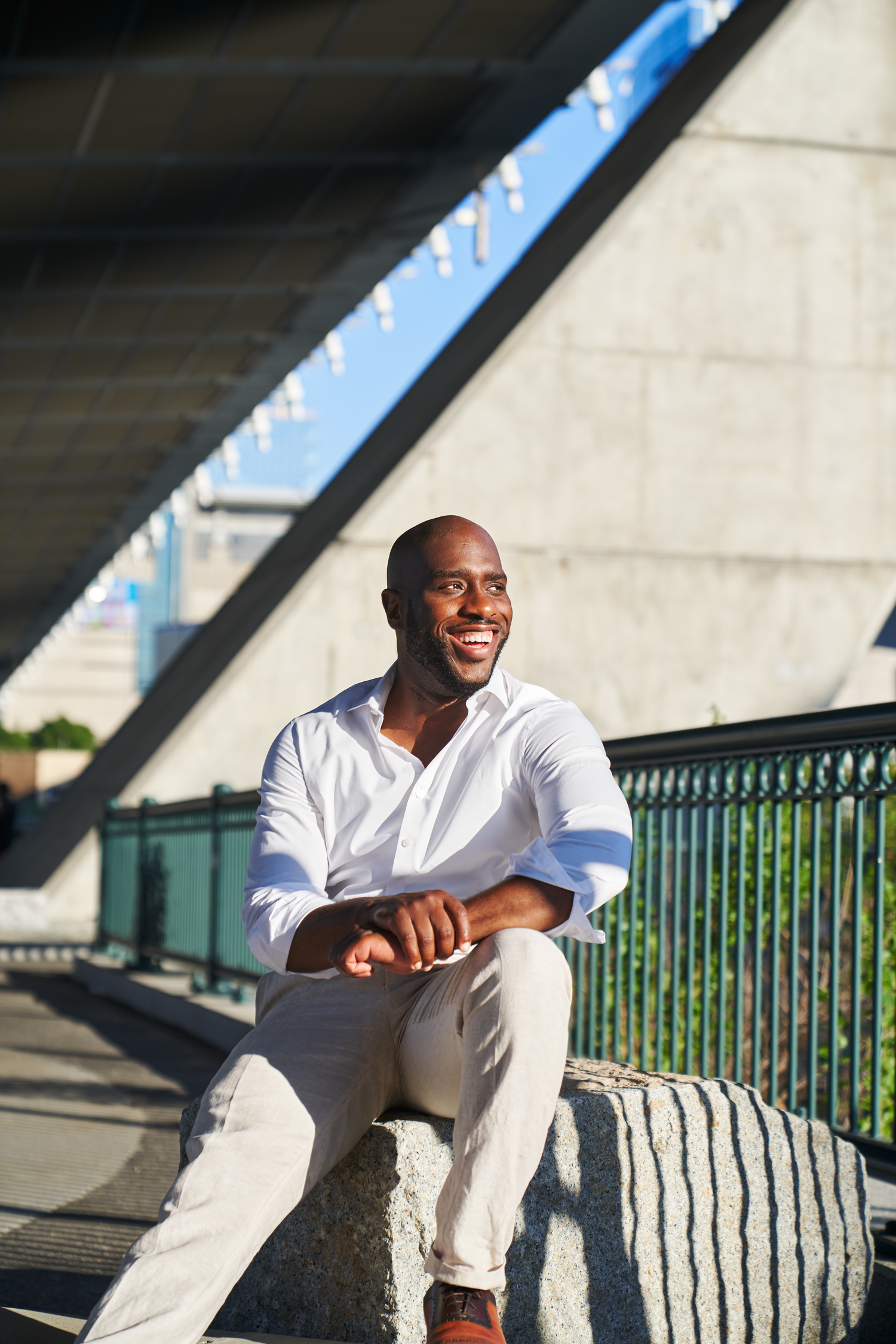 Smiling Black man in white shirt and khaki pants sitting on stone steps in an urban setting on a sunny day.