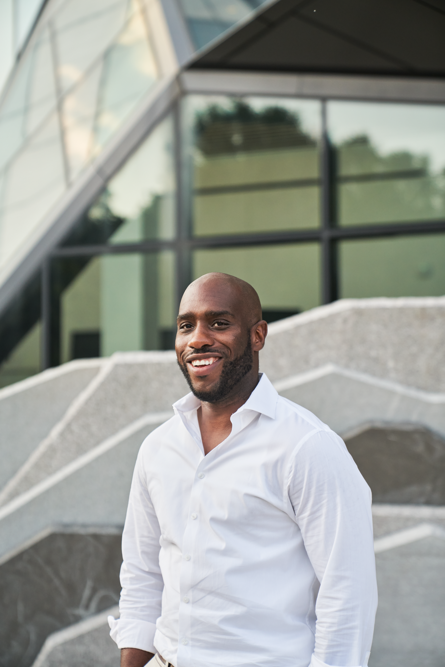 Smiling bald Black man with beard wearing white shirt standing outside modern glass building.