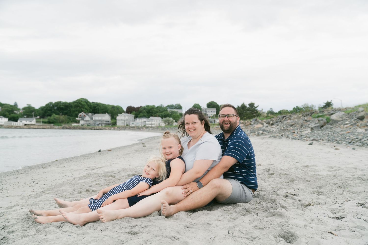 Family of four sitting together on a sandy beach, smiling, with a coastal town and cloudy sky in the background.