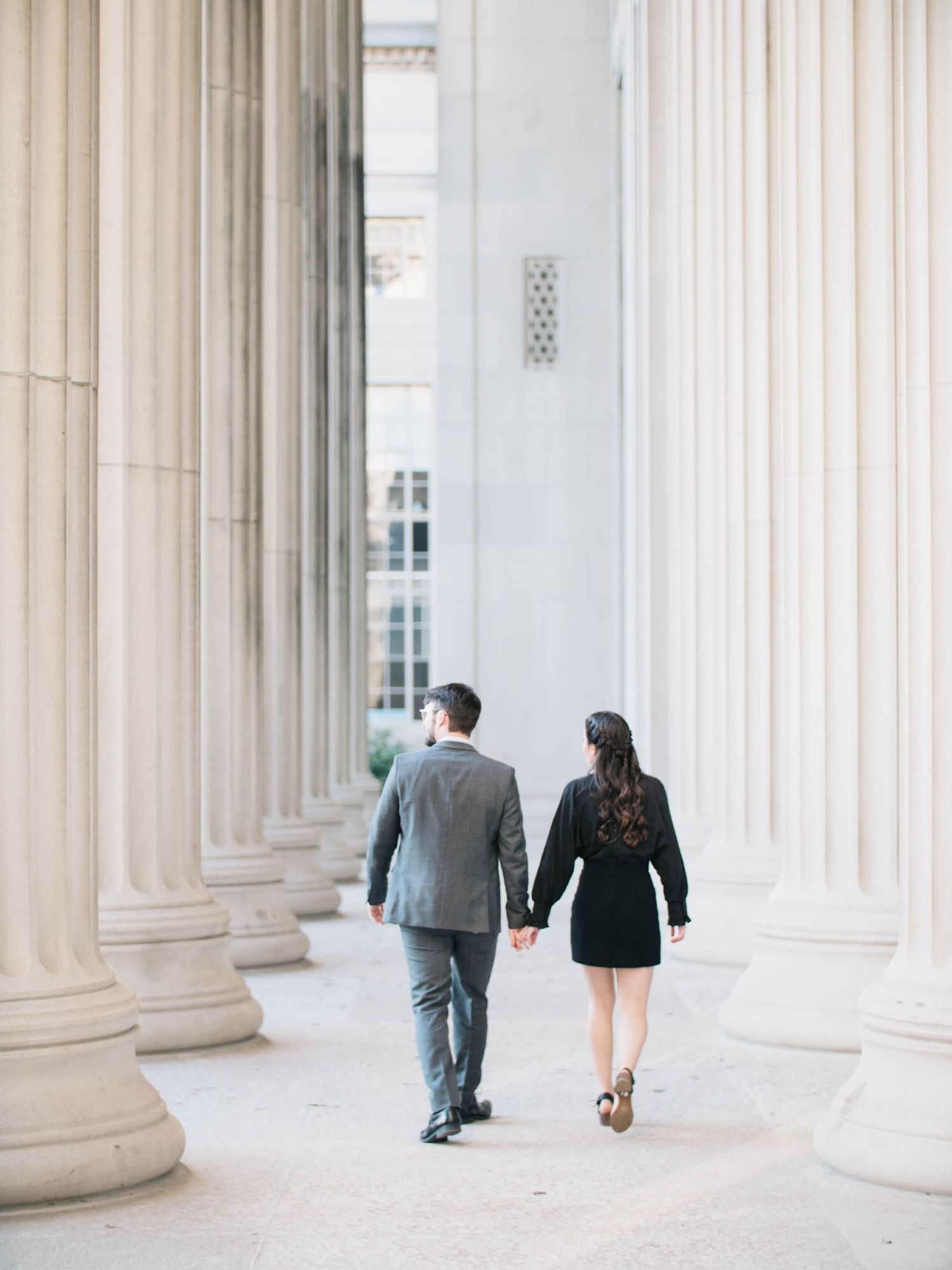 Couple holding hands walking between tall stone columns of a classical building exterior.
