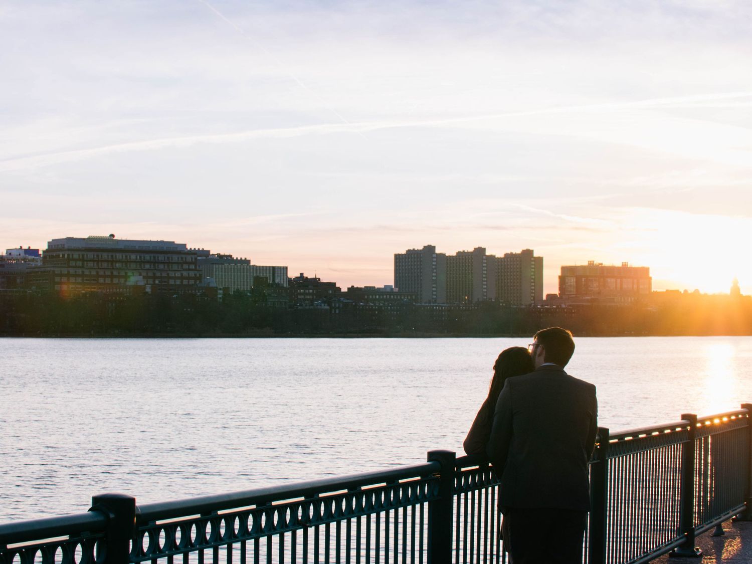 Couple standing together at waterfront railing watching a beautiful sunset over the city skyline.
