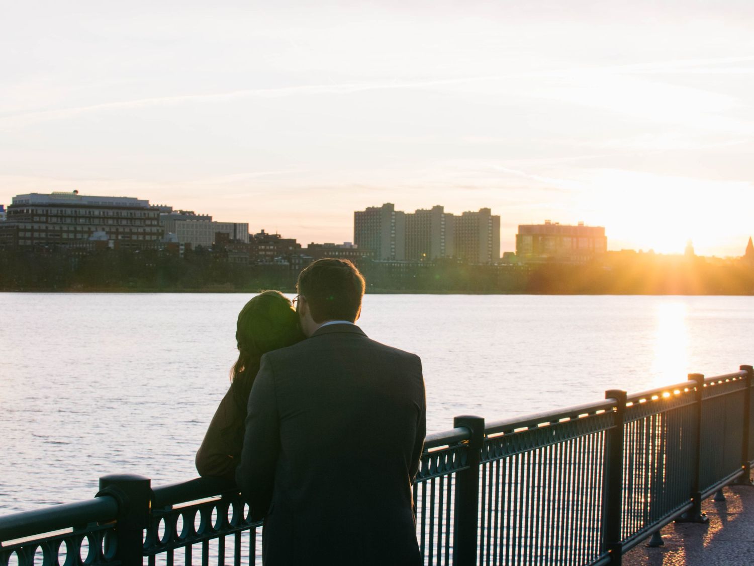 Couple embracing at sunset by a waterfront railing with Boston city skyline in background.