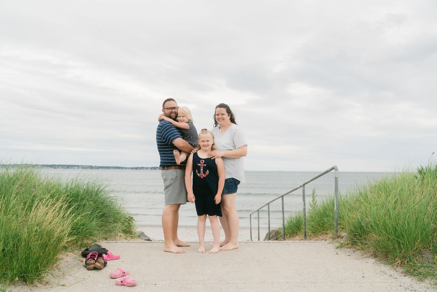 Family of four posing together on a sandy beach path with ocean views on a cloudy day.