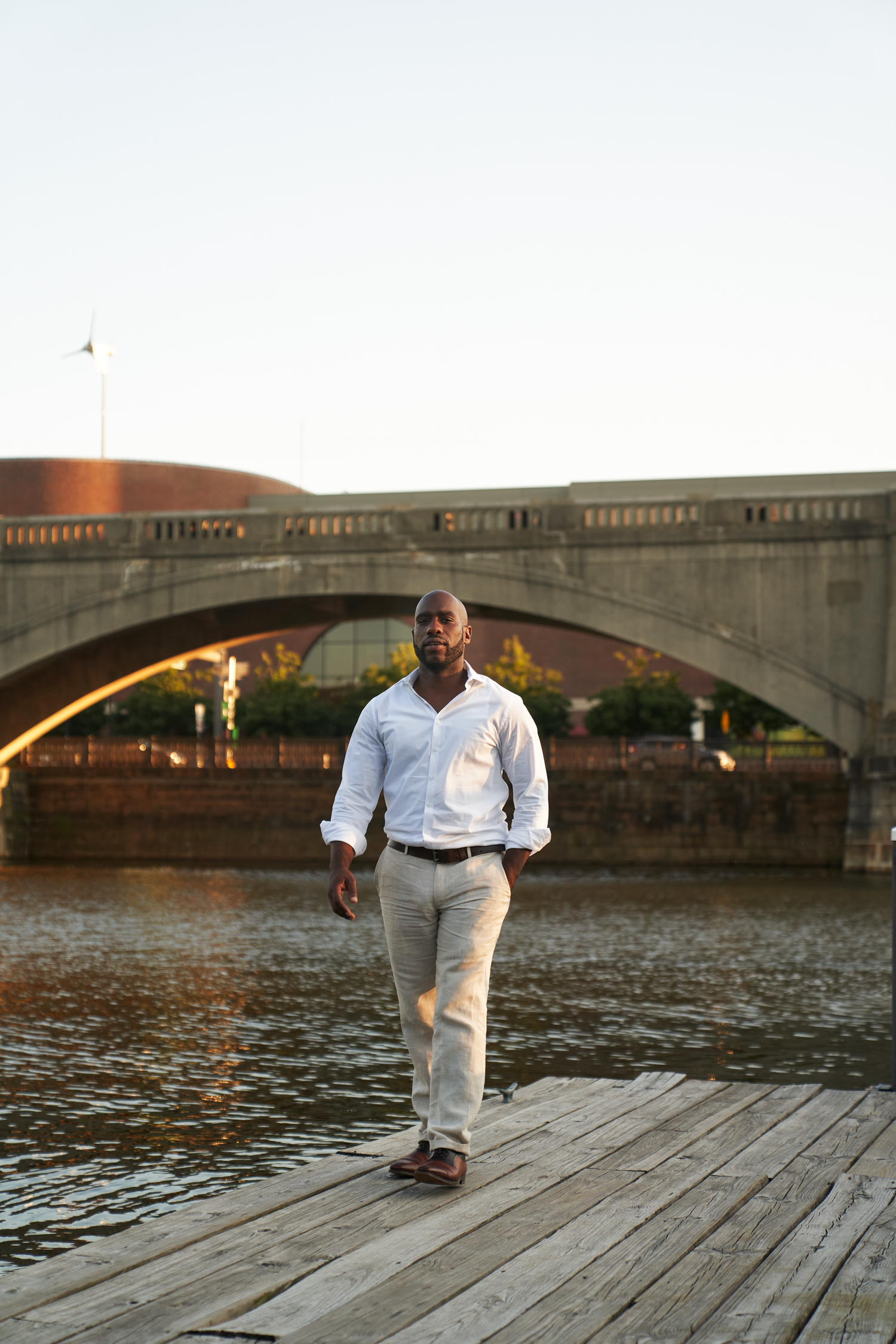 A confident Black man in a white shirt and khaki pants stands on a wooden dock with an arched bridge over a river behind him.