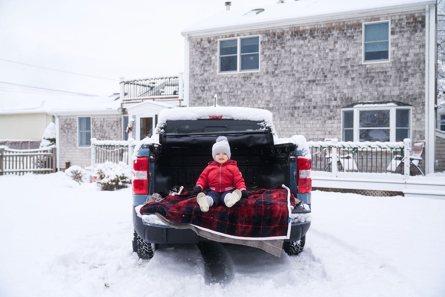 Baby in red snowsuit sitting alone on plaid blanket in snowy truck bed outdoors.