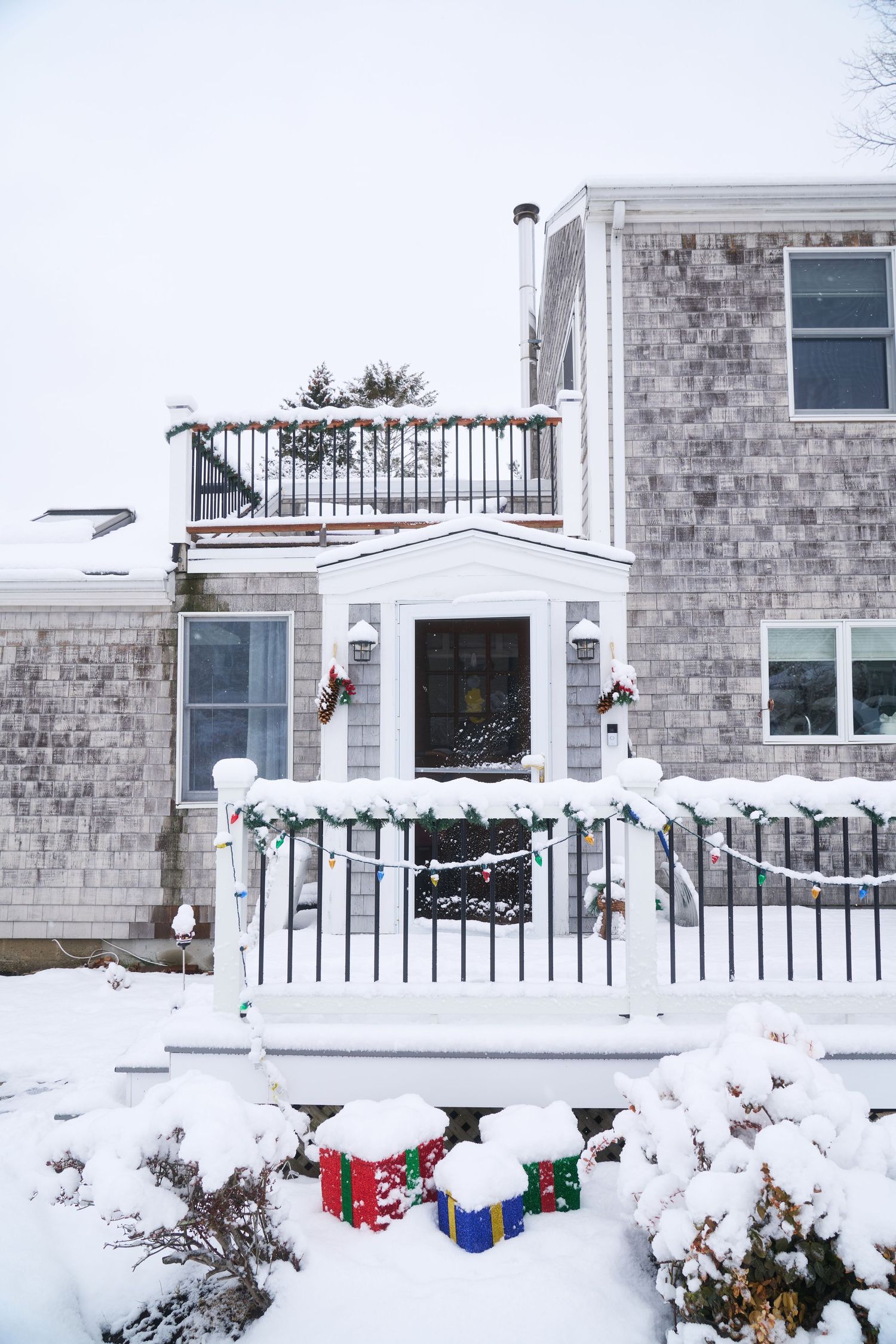 Snow-covered New England style home decorated with Christmas wreaths and colorful gift boxes outside.