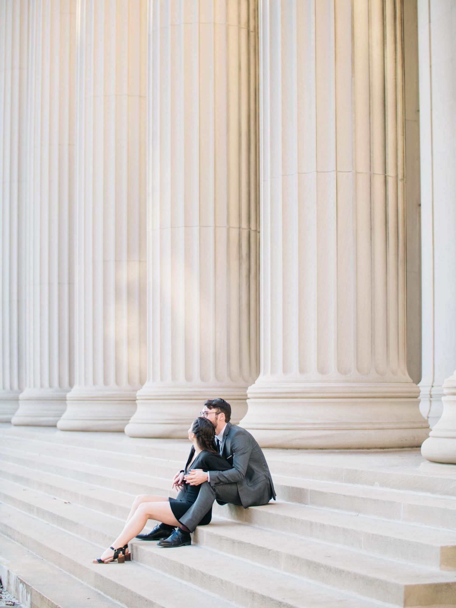 Couple sitting closely on steps sharing a kiss beside tall classical stone columns.