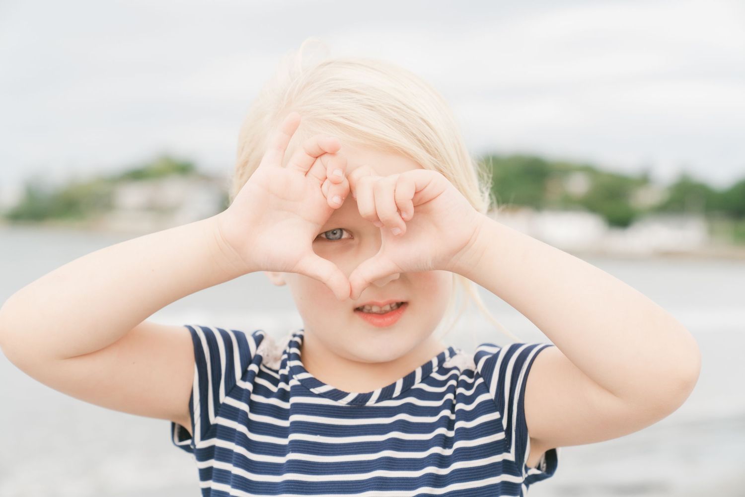 Cute blonde child making a heart shape with hands over eyes at the beach.