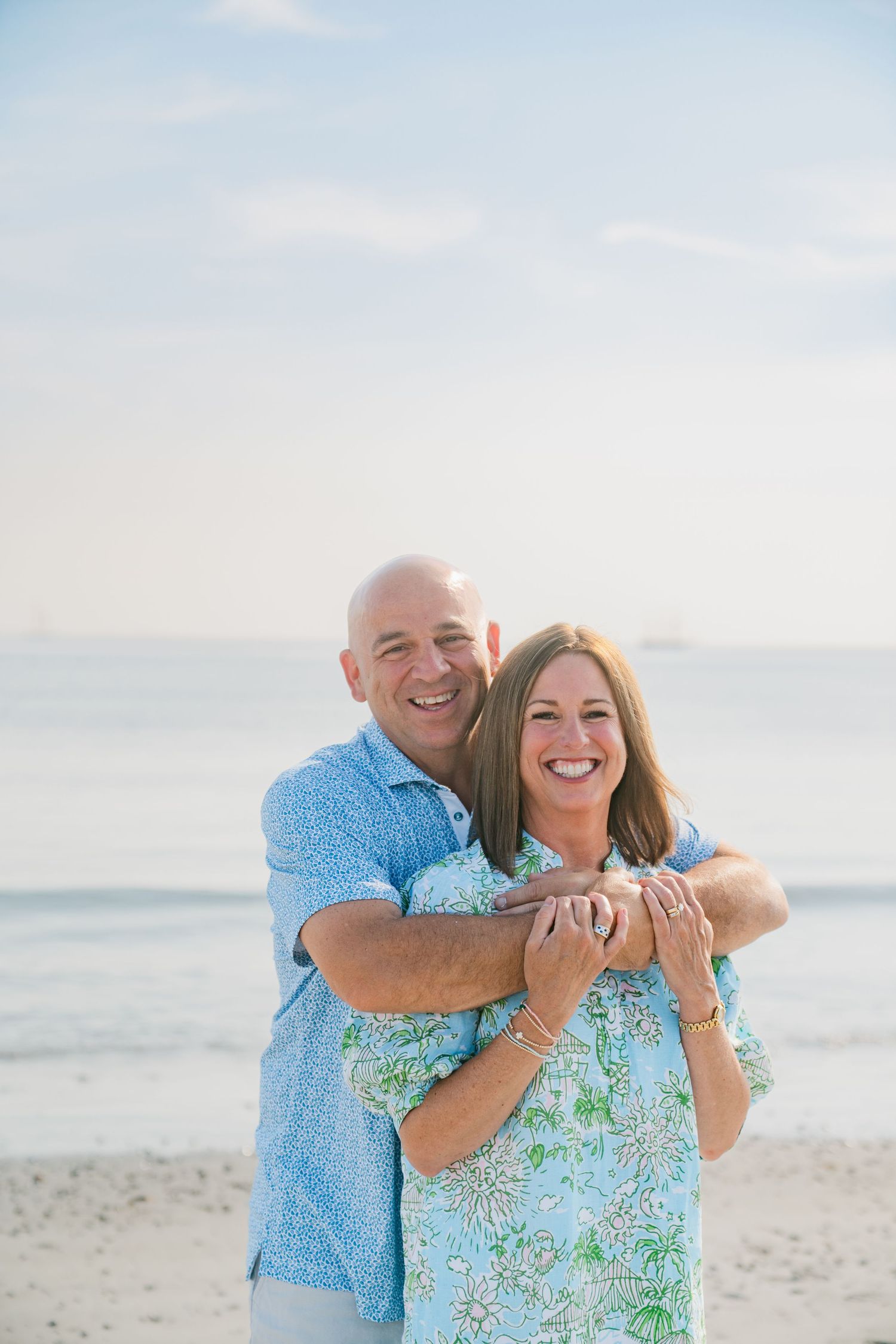 A smiling couple embraces on a sunny beach, man in blue shirt hugging woman in floral top with ocean in background.