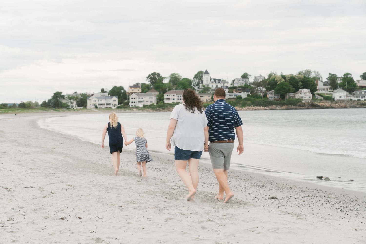 Family of four walking barefoot on a sandy beach with coastal homes in the background on a cloudy day.