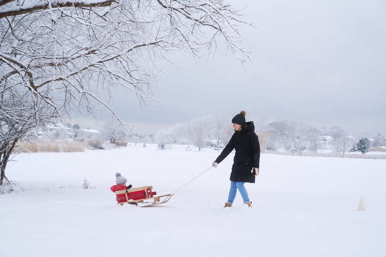 Woman in black coat pulling a young child on a red wooden sled through a snowy landscape.