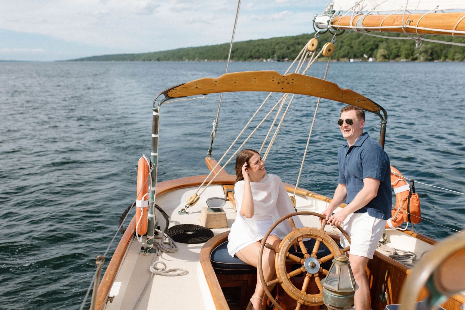 Couple enjoying a sunny sail on a classic wooden boat with a ship wheel on calm blue waters.
