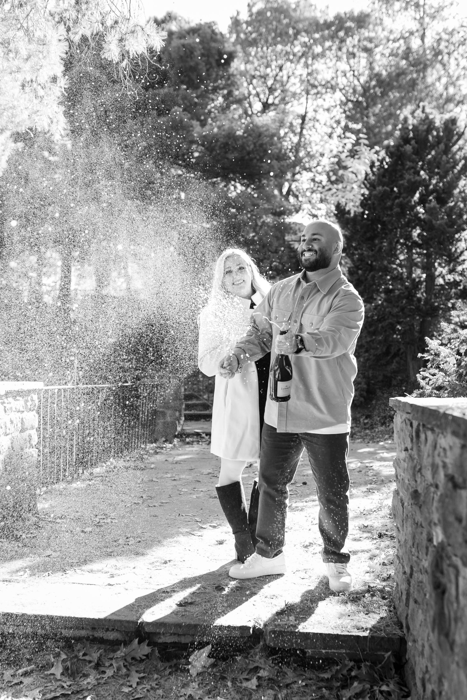 Black and white photo of a couple celebrating outdoors, man popping champagne bottle with spray of bubbles in a park setting.