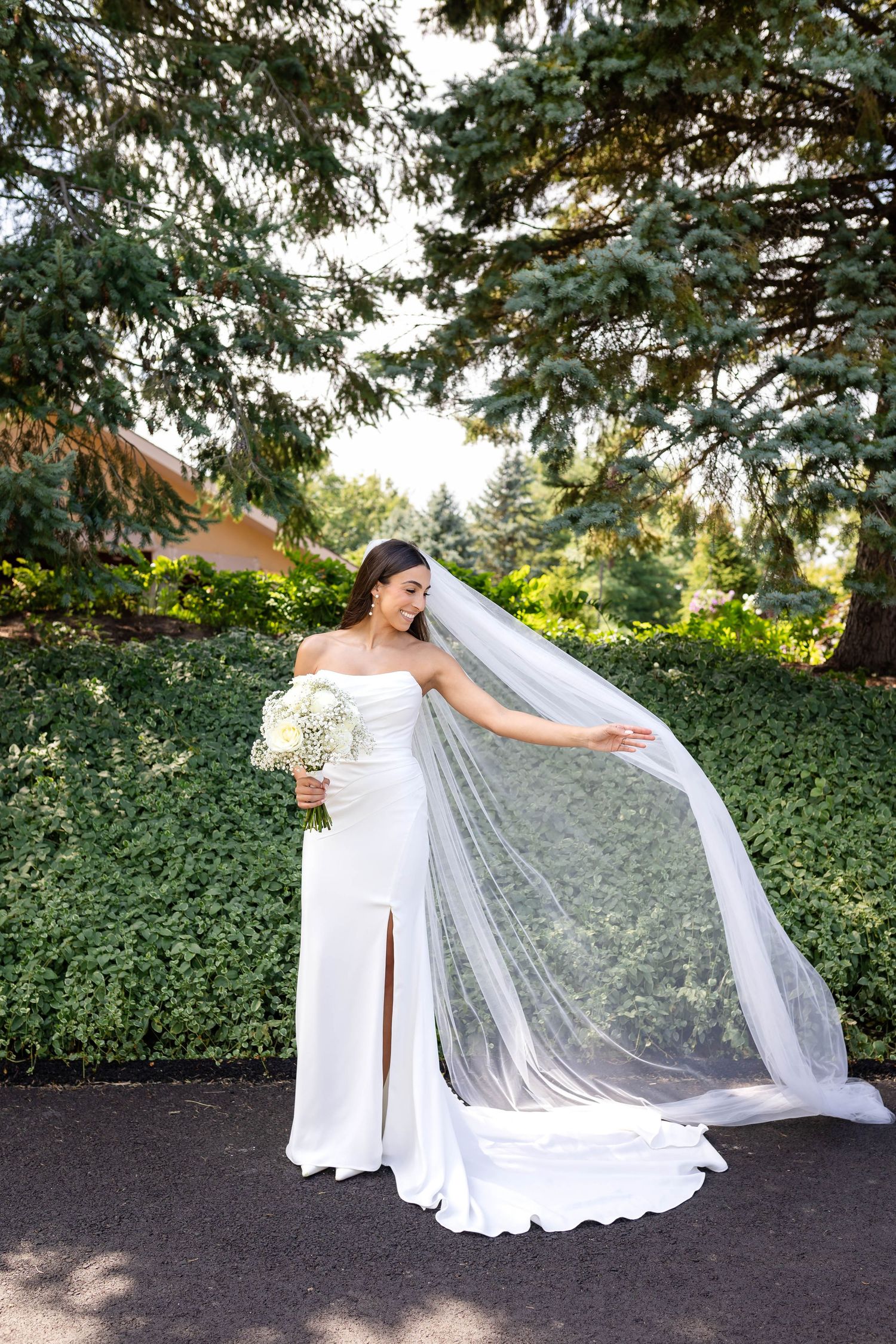 Bride in white strapless gown with flowing veil holds bouquet outdoors among lush green trees and hedges.