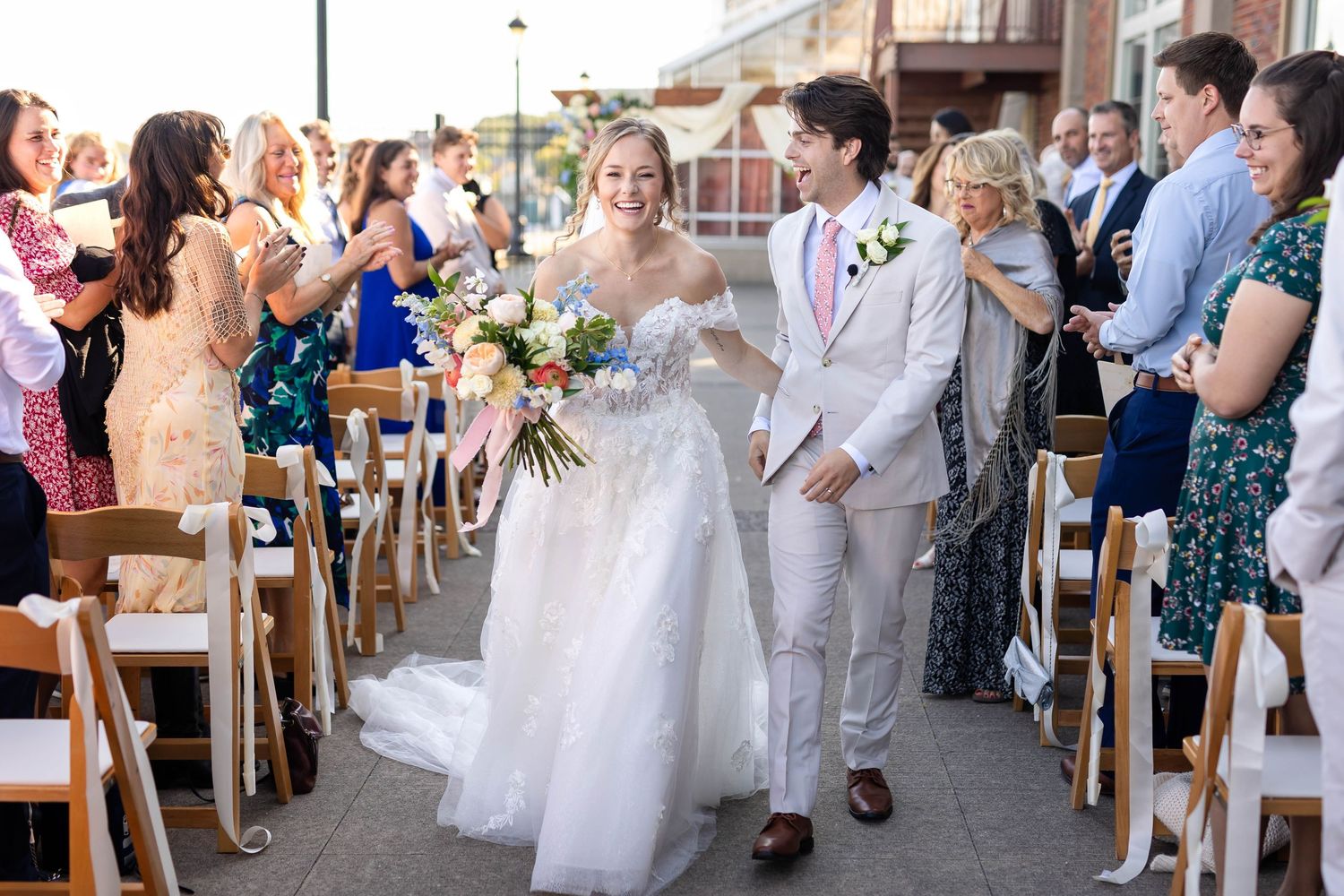 Wedding couple walks down aisle while guests celebrate during outdoor ceremony with wooden chairs and floral decor.