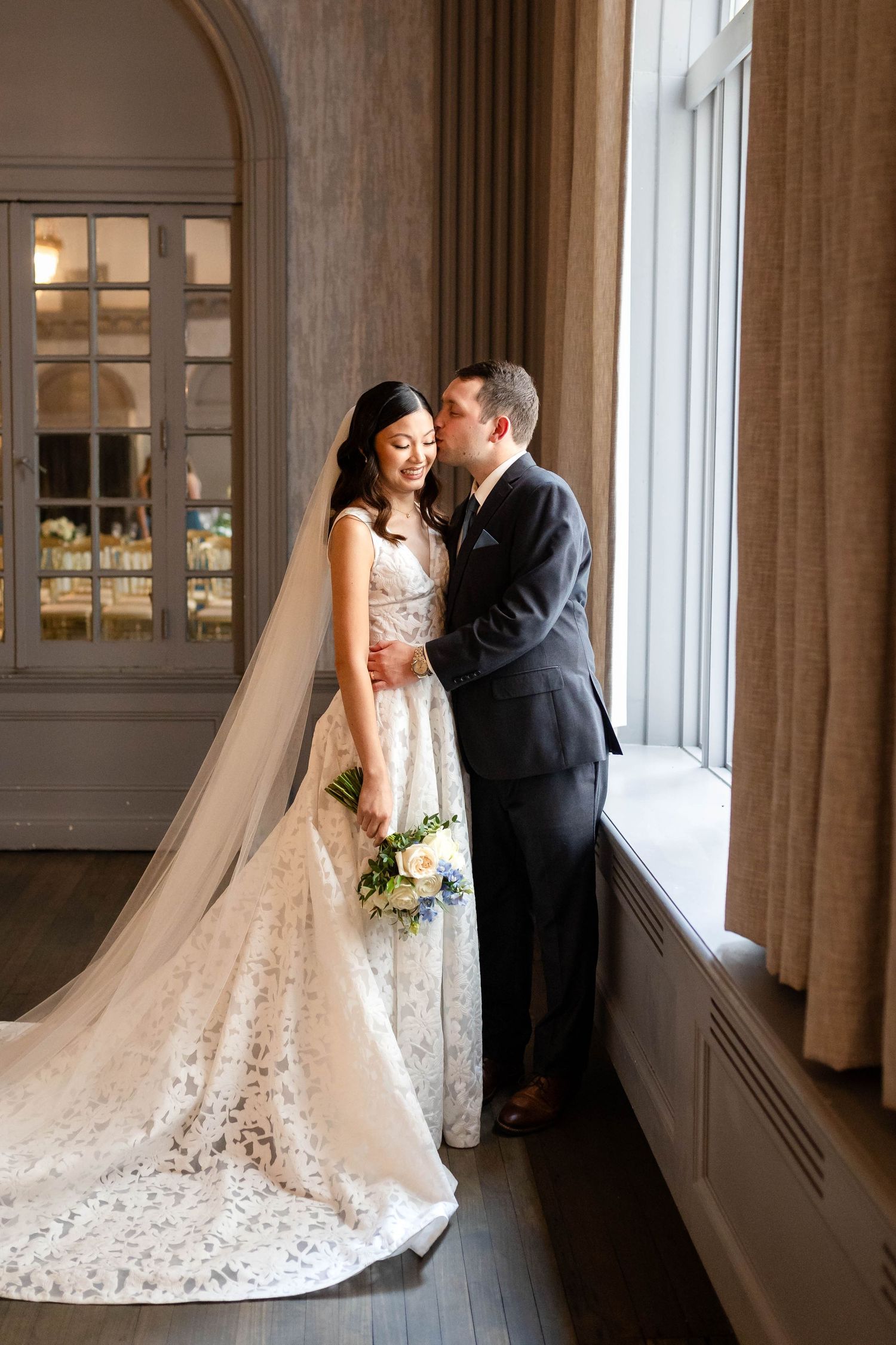 Bride in lace gown with veil and groom in dark suit share intimate moment by window holding blue floral bouquet.