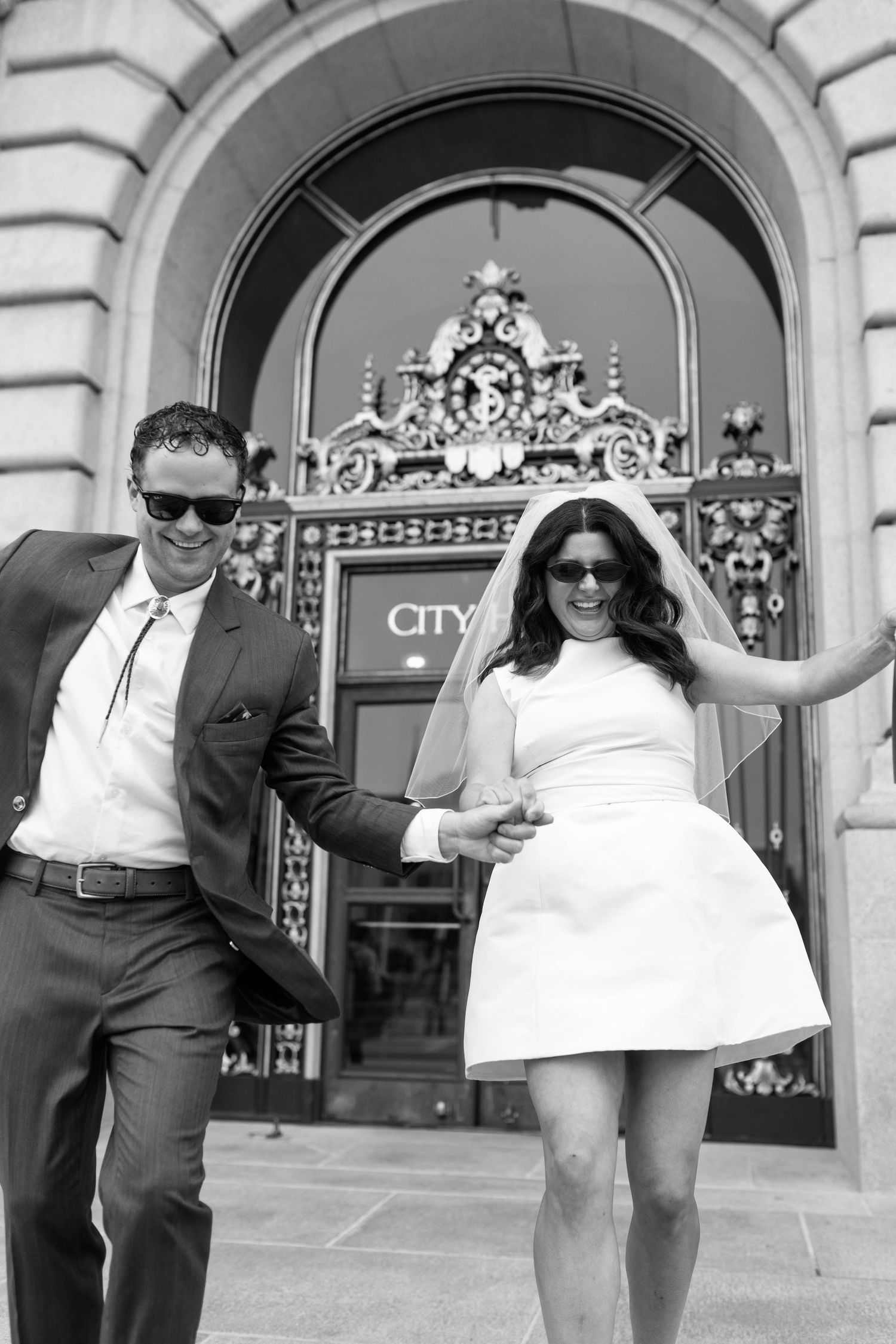 Black and white photo of a newlywed couple in sunglasses holding hands outside City Hall.