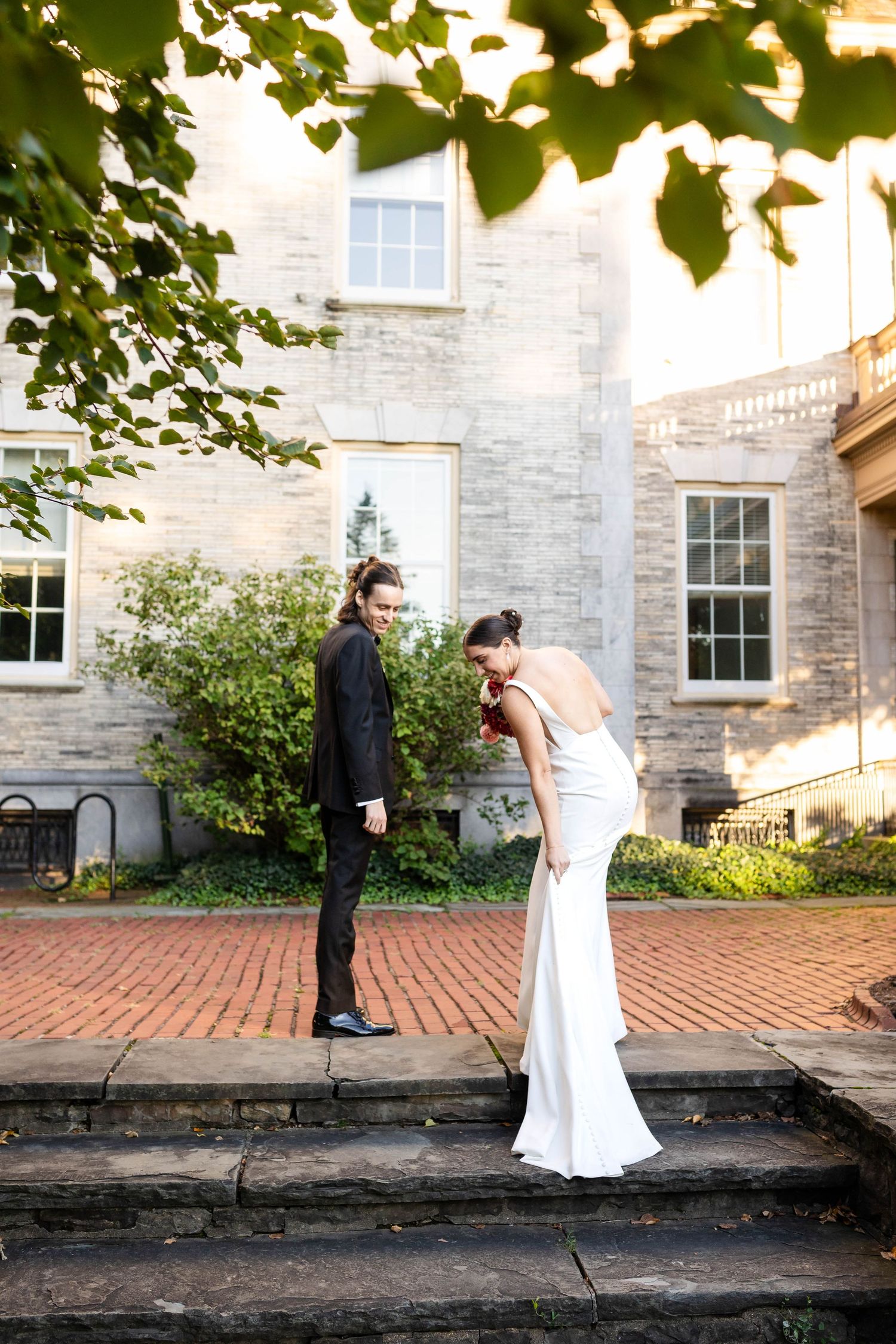 Bride in white gown and groom in black suit share a romantic moment on brick steps outside a stone building.