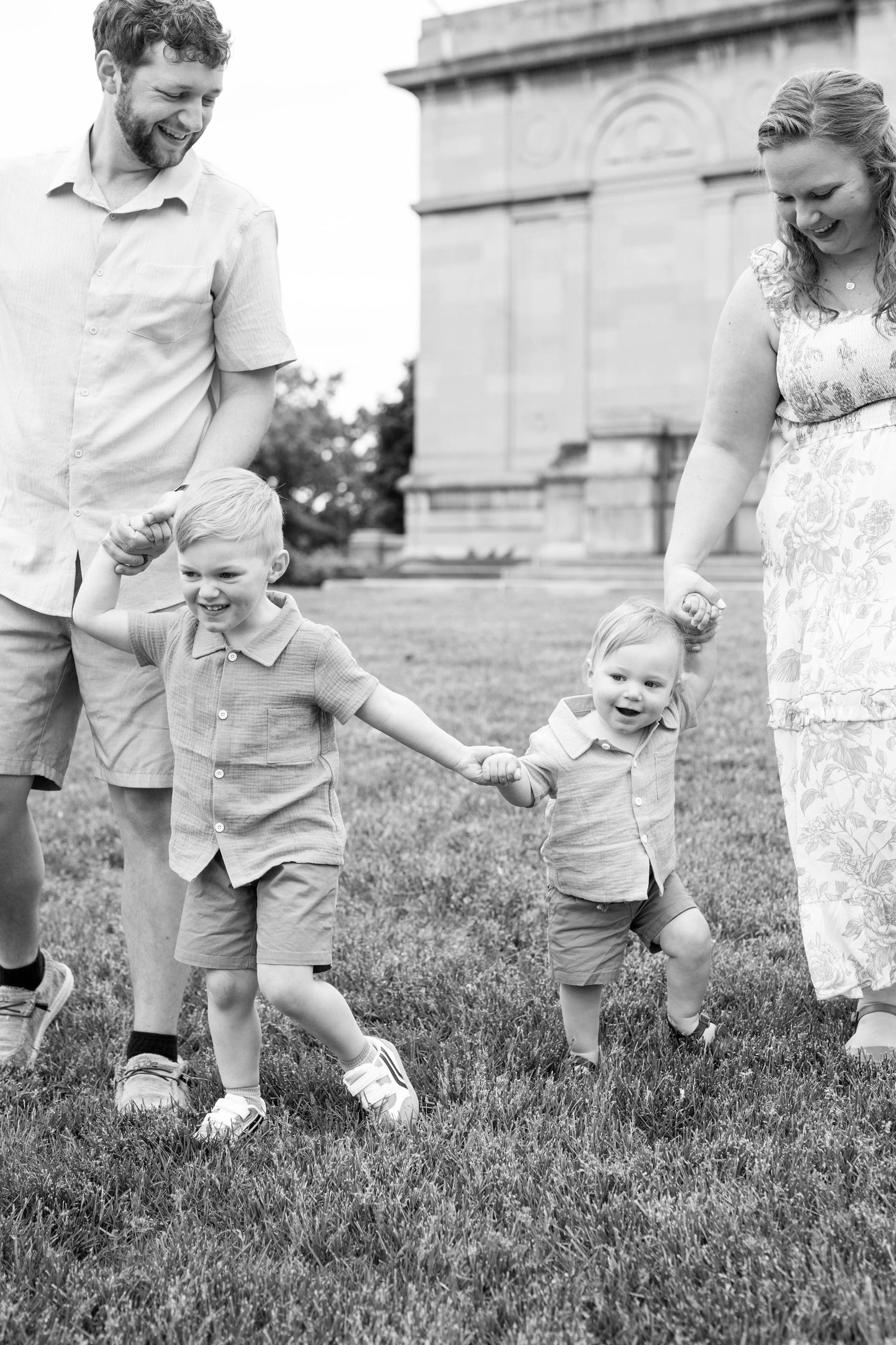 Black and white photo of a happy family with two young boys holding hands and walking on grass near a historic building.