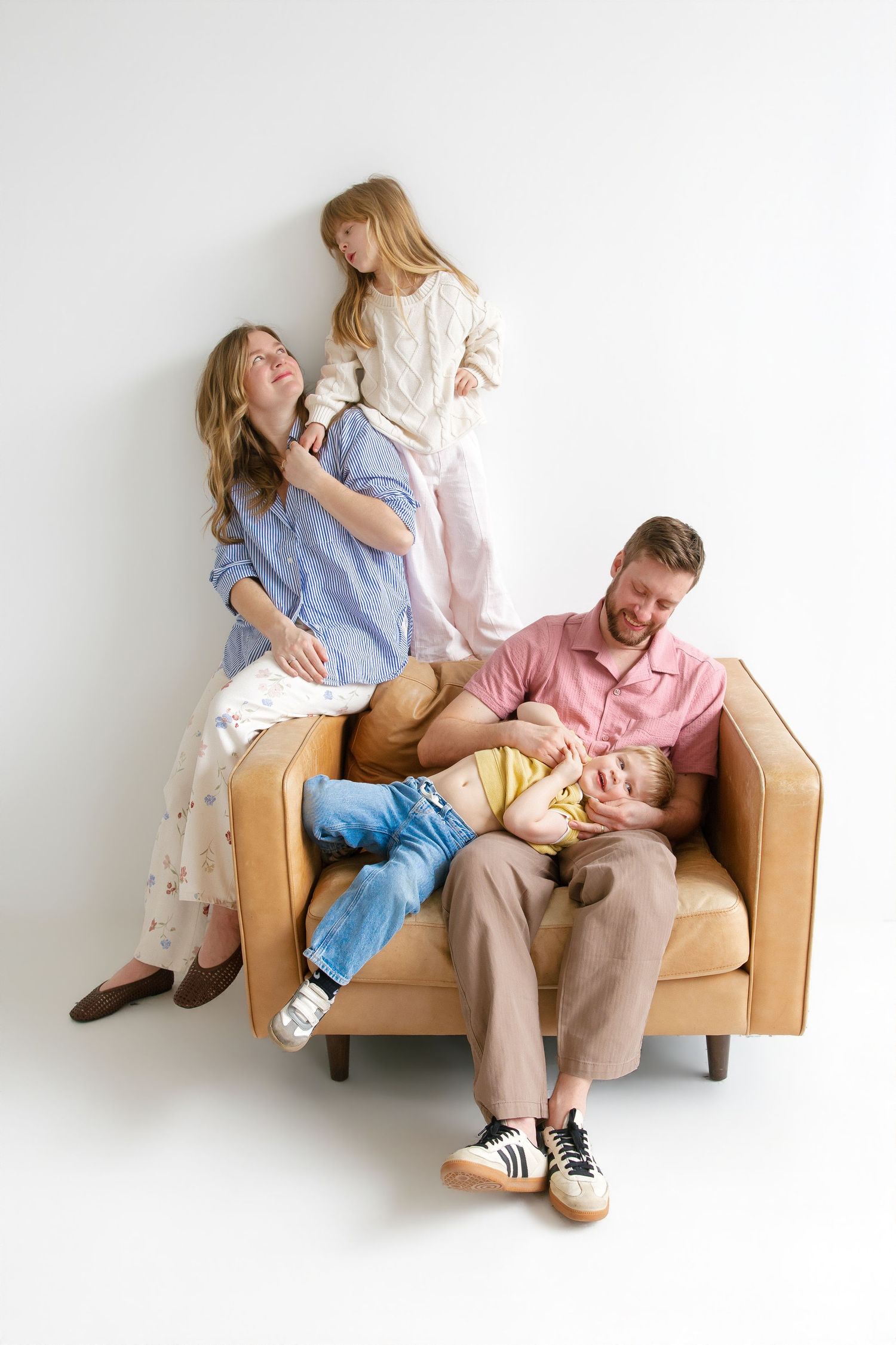 A joyful family of four relaxing together on a tan leather armchair against a white wall.