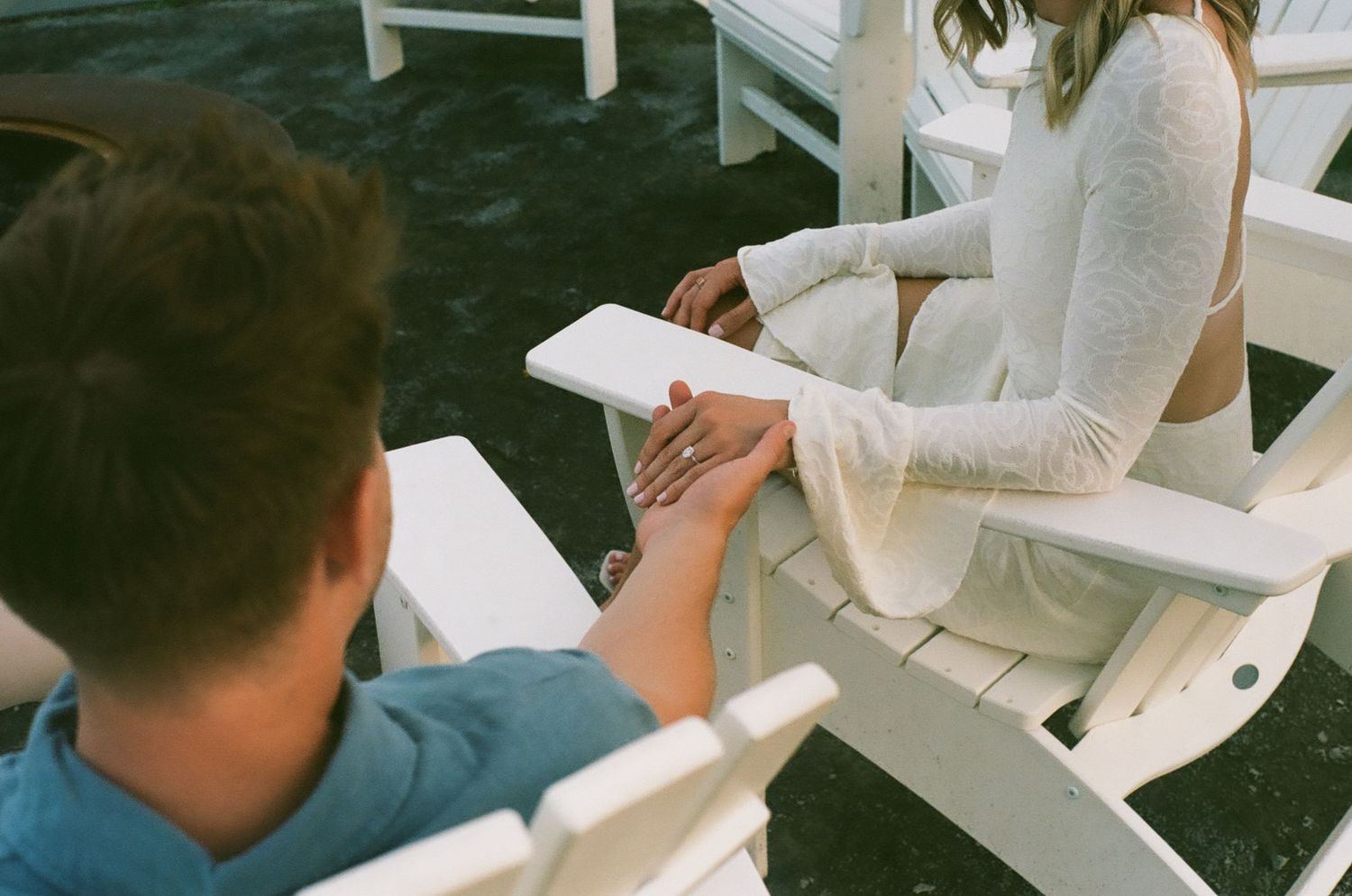Couple holding hands while sitting in white Adirondack chairs outdoors, woman in white lace dress.