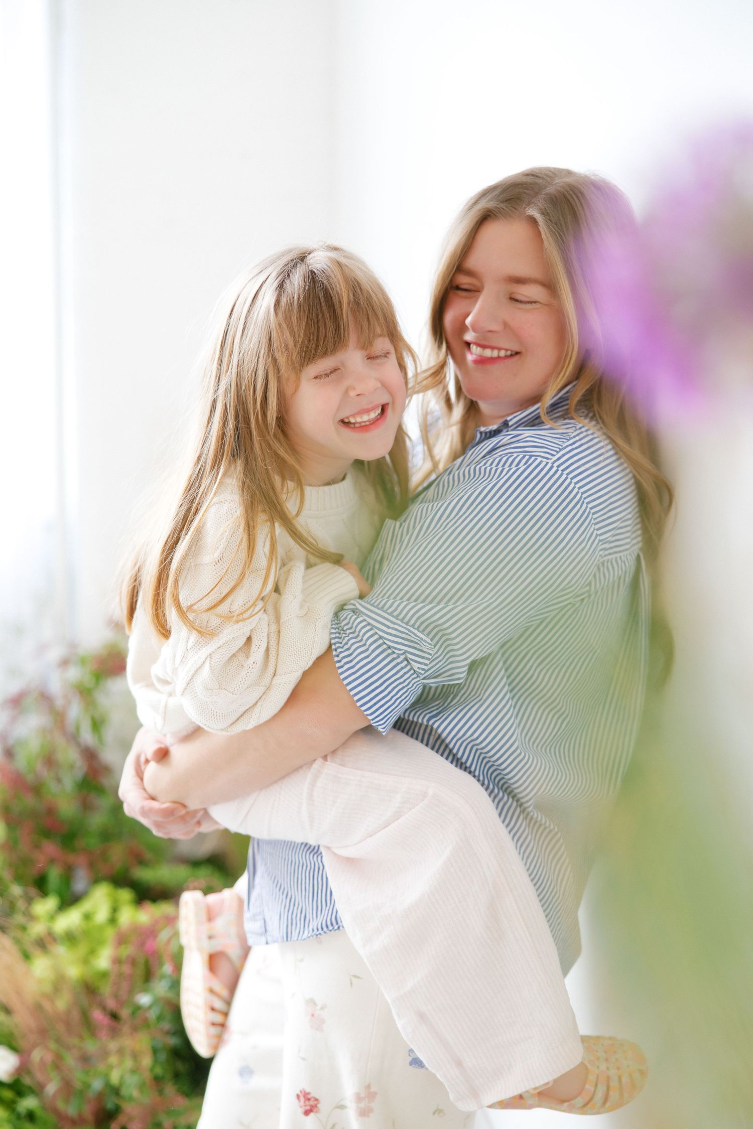 A woman and young girl share a joyful embrace, both laughing, surrounded by colorful flowers in bright light.