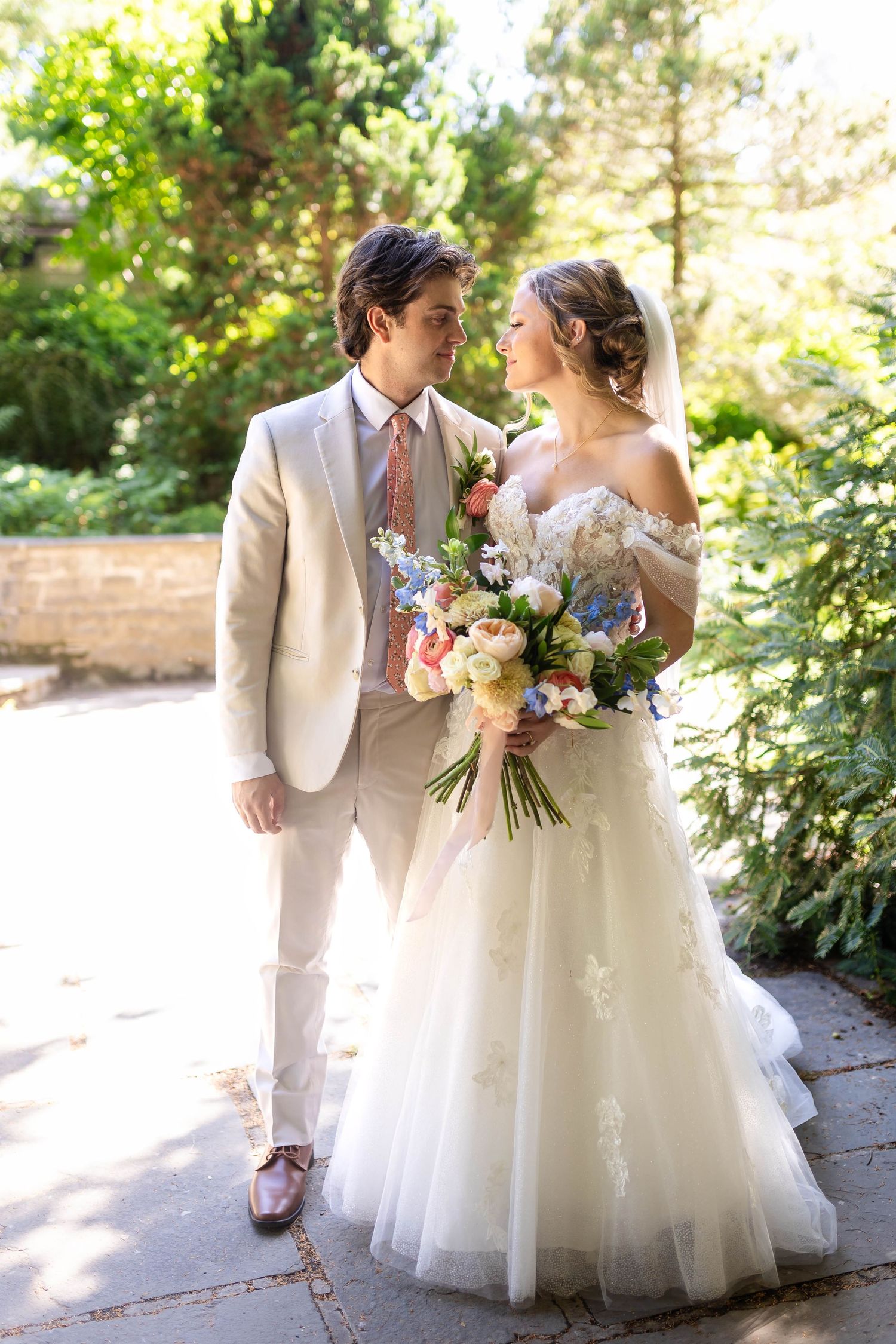 A bride and groom share a romantic moment outdoors, she holds a colorful bouquet, he wears a beige suit with a pink tie.