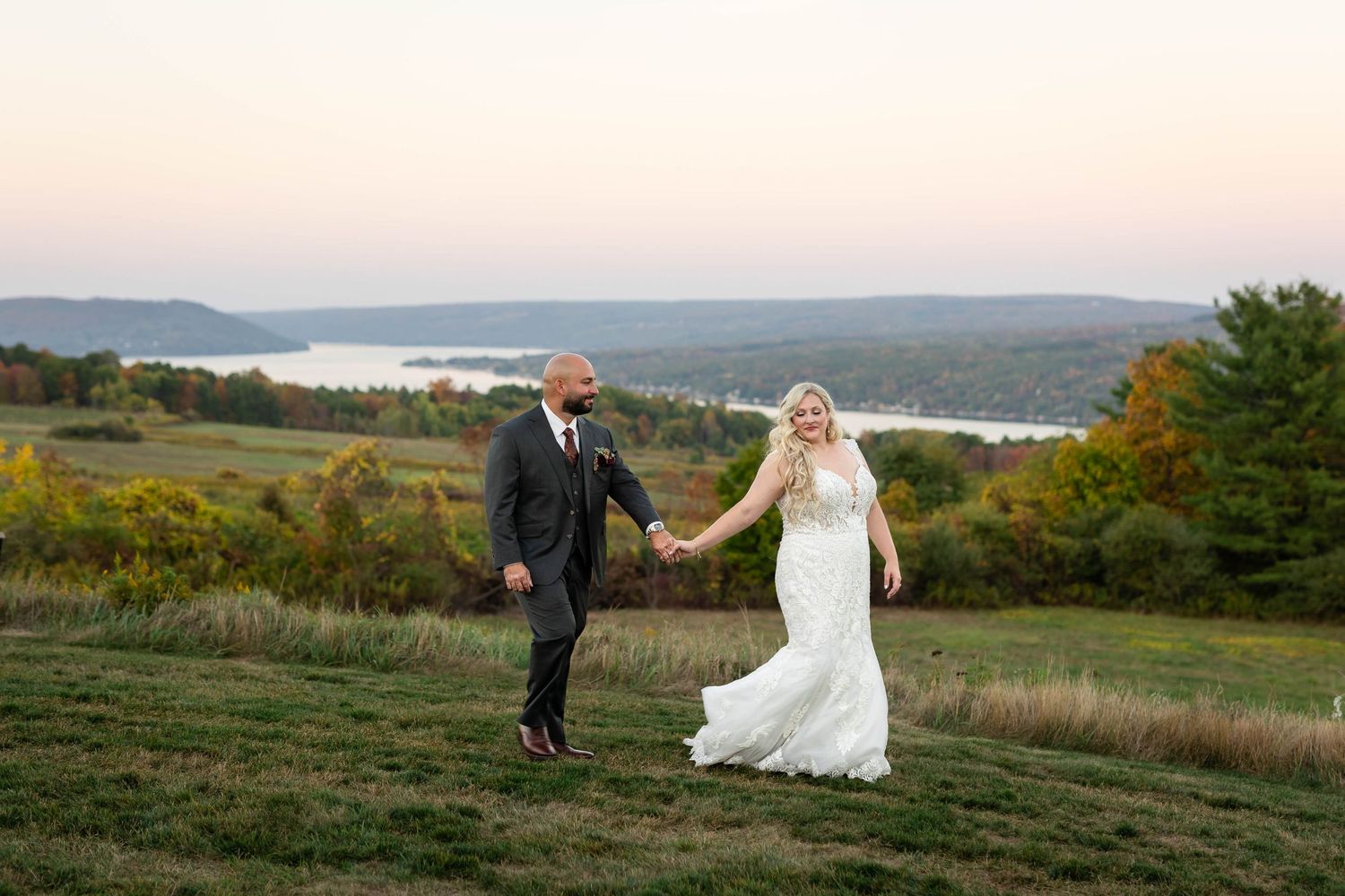 A couple in wedding attire walks through scenic fields overlooking a lake during sunset.