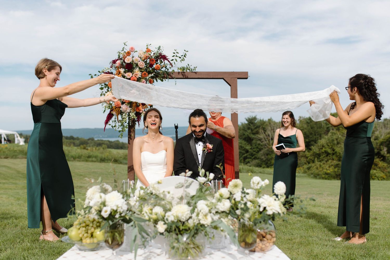 A wedding party celebrates on a grassy hilltop with bridesmaids in emerald green dresses and floral bouquets.