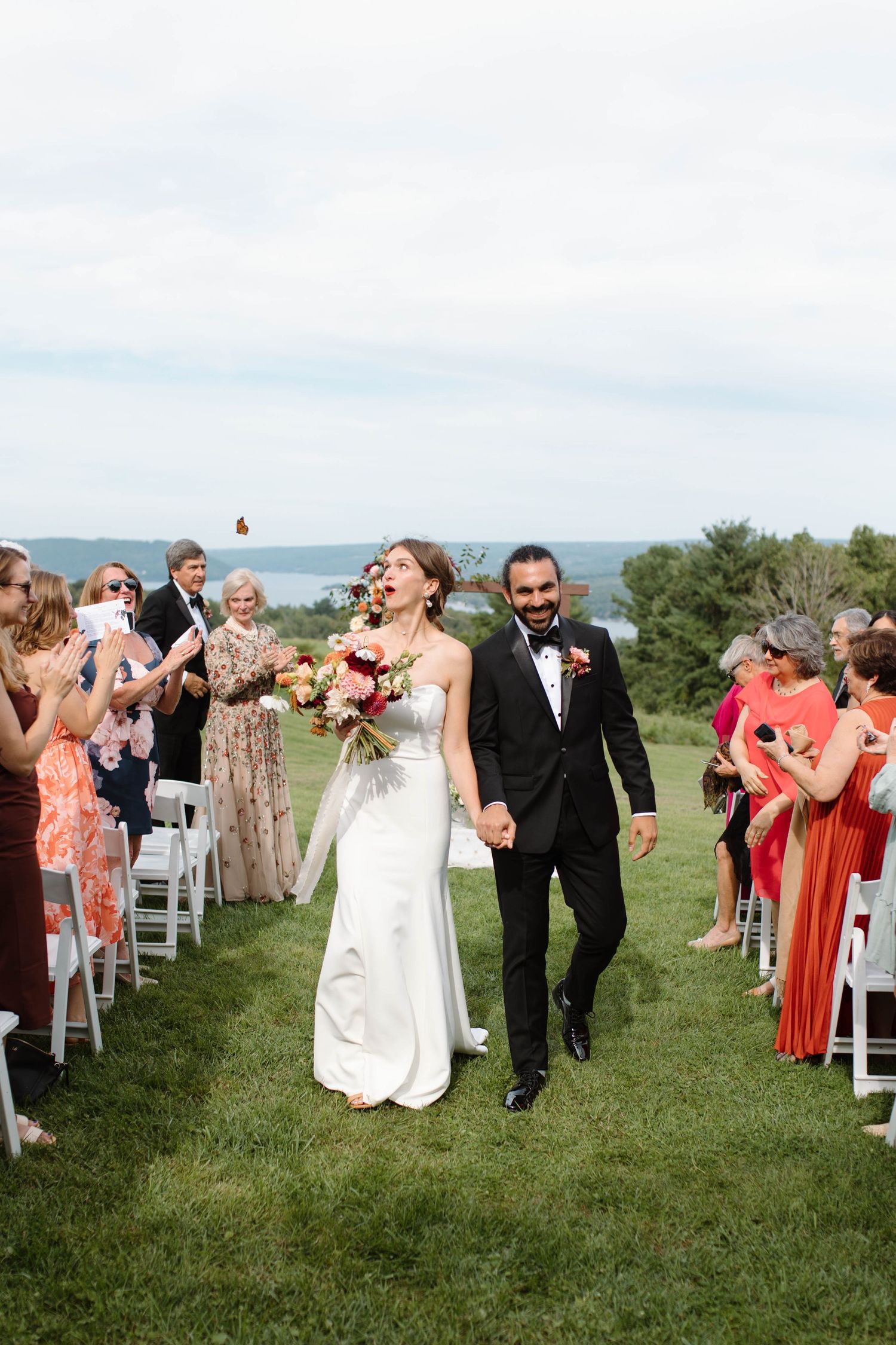 Joyful newlyweds walk down outdoor aisle holding hands, bride in white gown with bouquet, guests cheering with water views.
