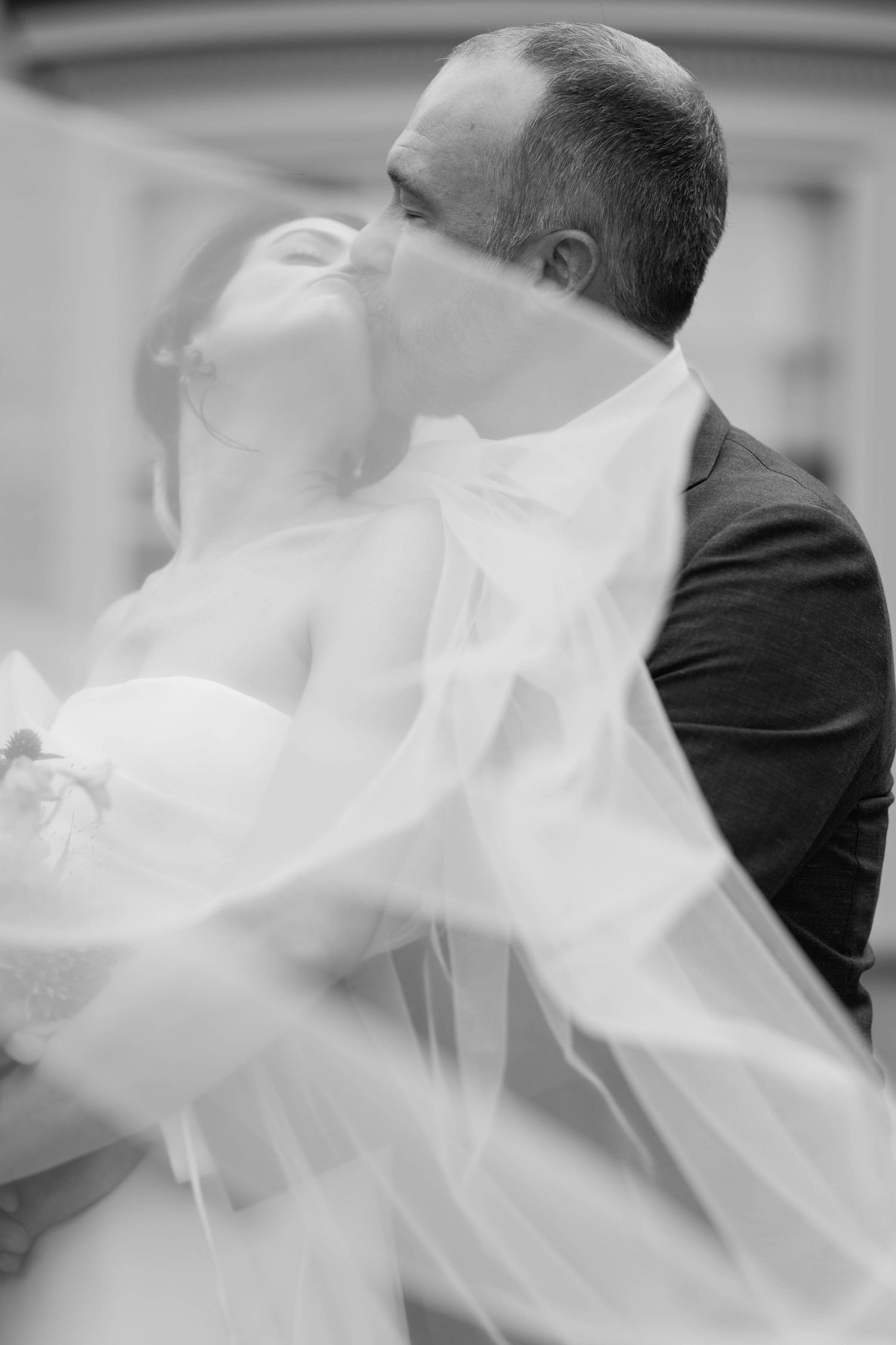 Black and white photo of a bride and groom sharing a kiss under flowing wedding veil.
