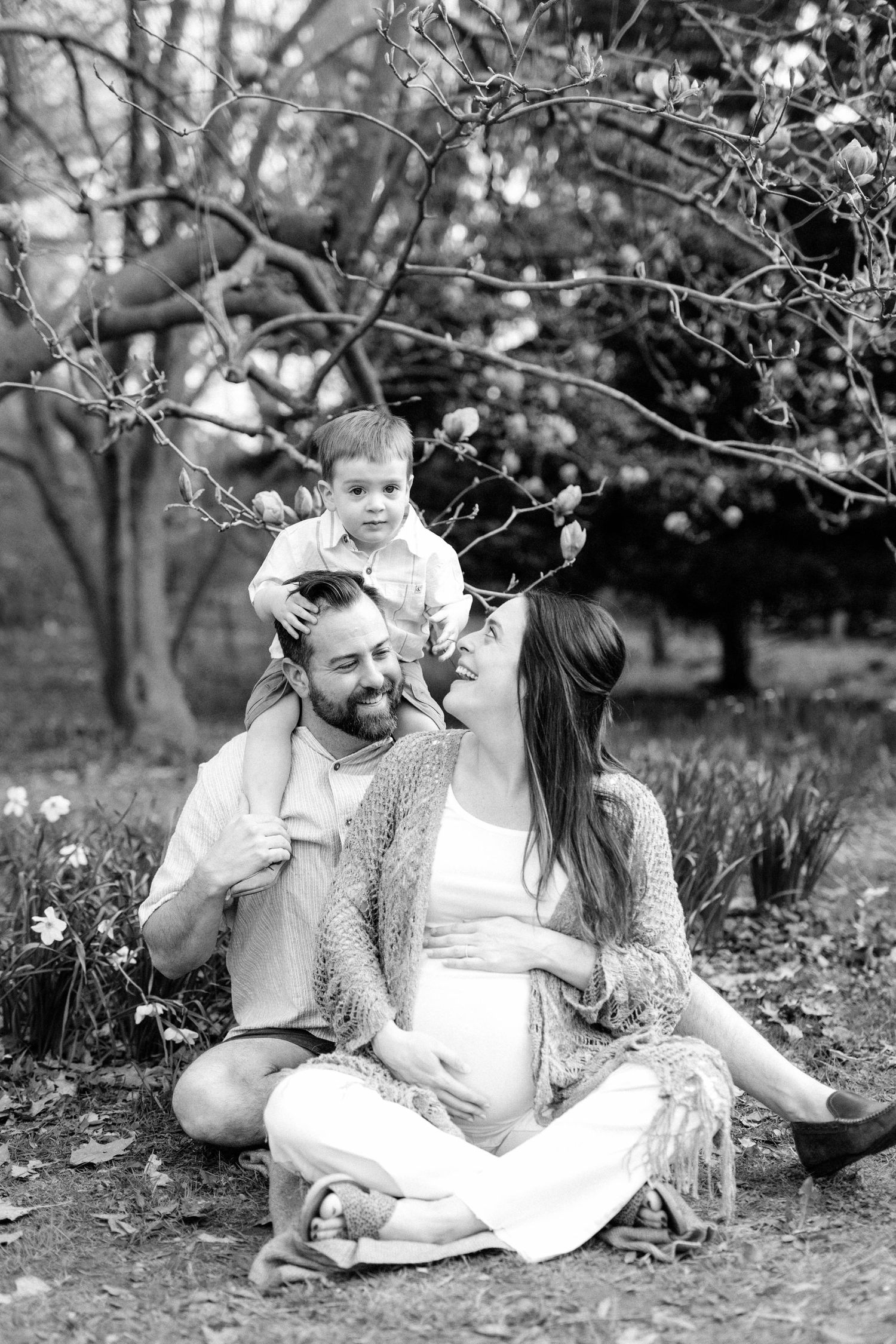 Black and white photo of a pregnant woman, man, and toddler sitting together outdoors under blooming trees.