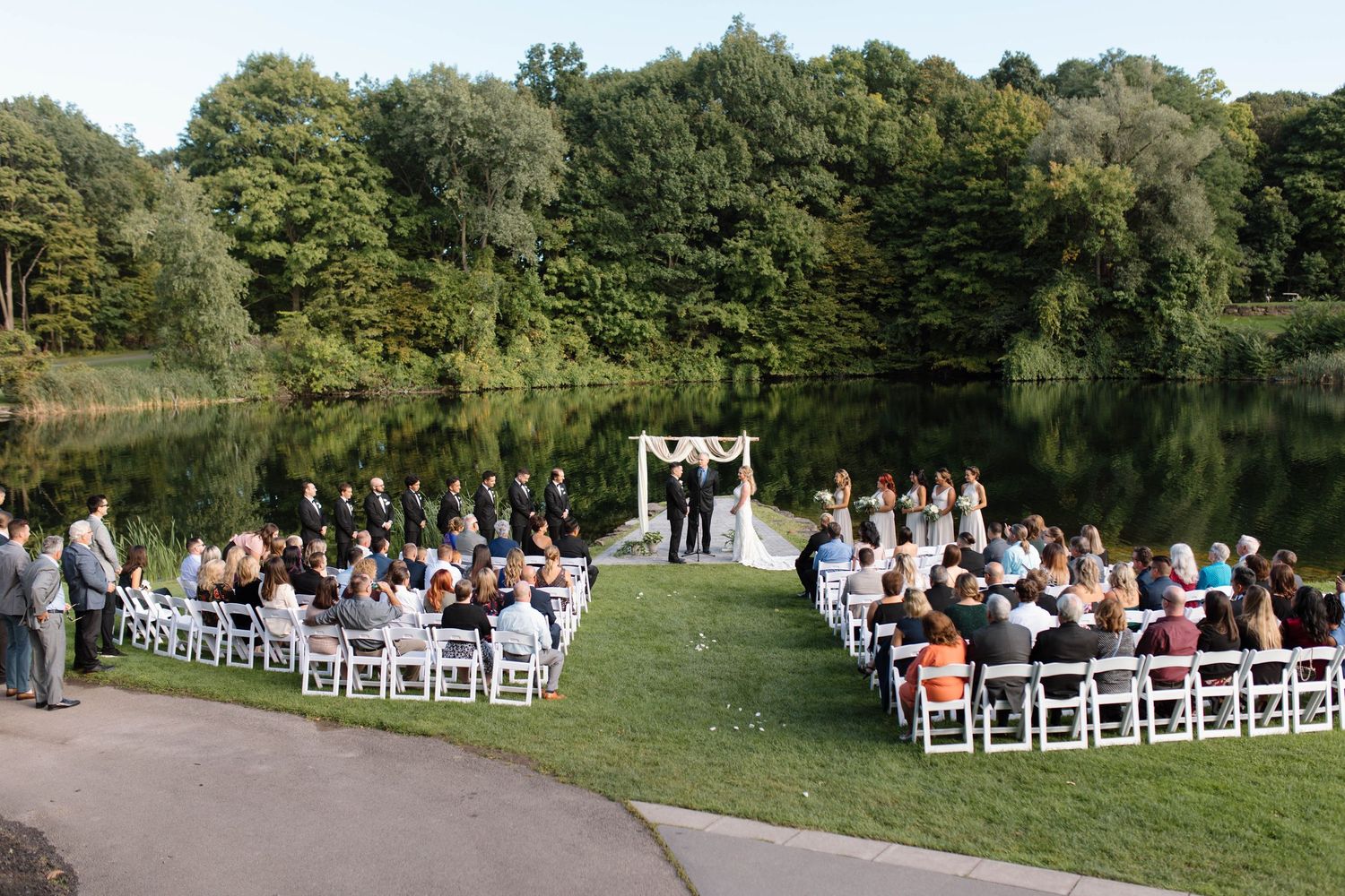 Outdoor wedding ceremony setup with white chairs arranged in rows on a lush green lawn near a serene lake.