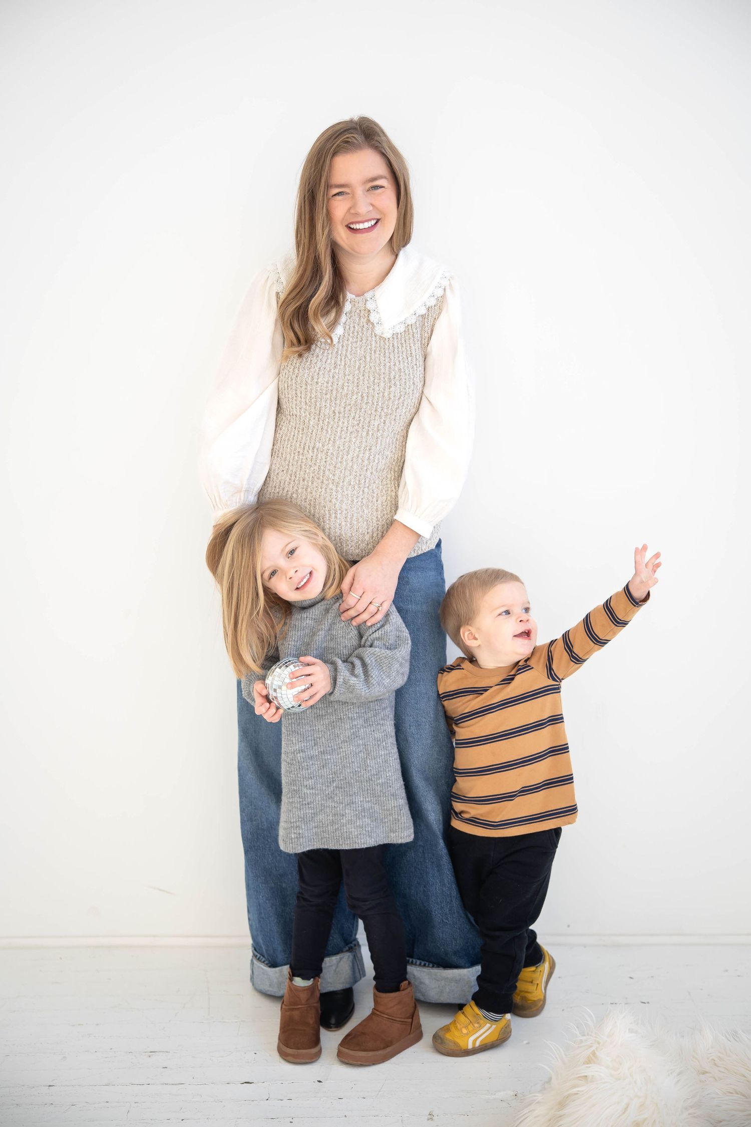 A smiling woman in a knit vest poses with two young children against a white wall in a cozy family portrait.