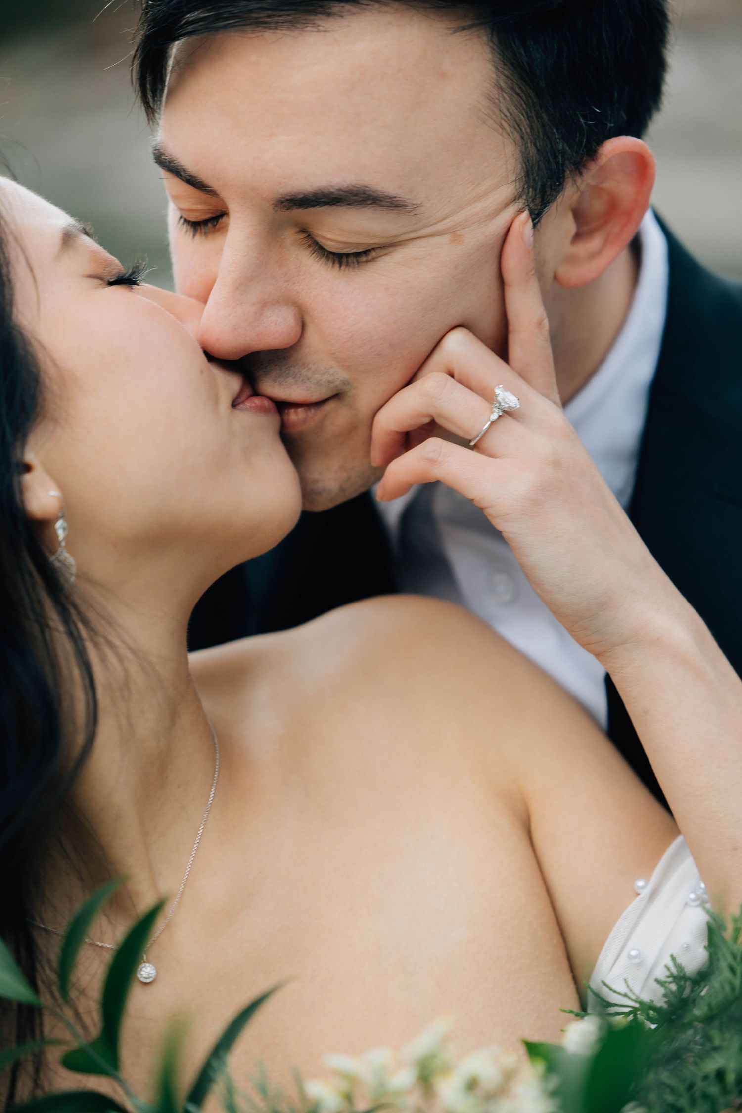 Bride and groom sharing a kiss, bride wearing diamond engagement ring, holding greenery bouquet, close-up portrait.