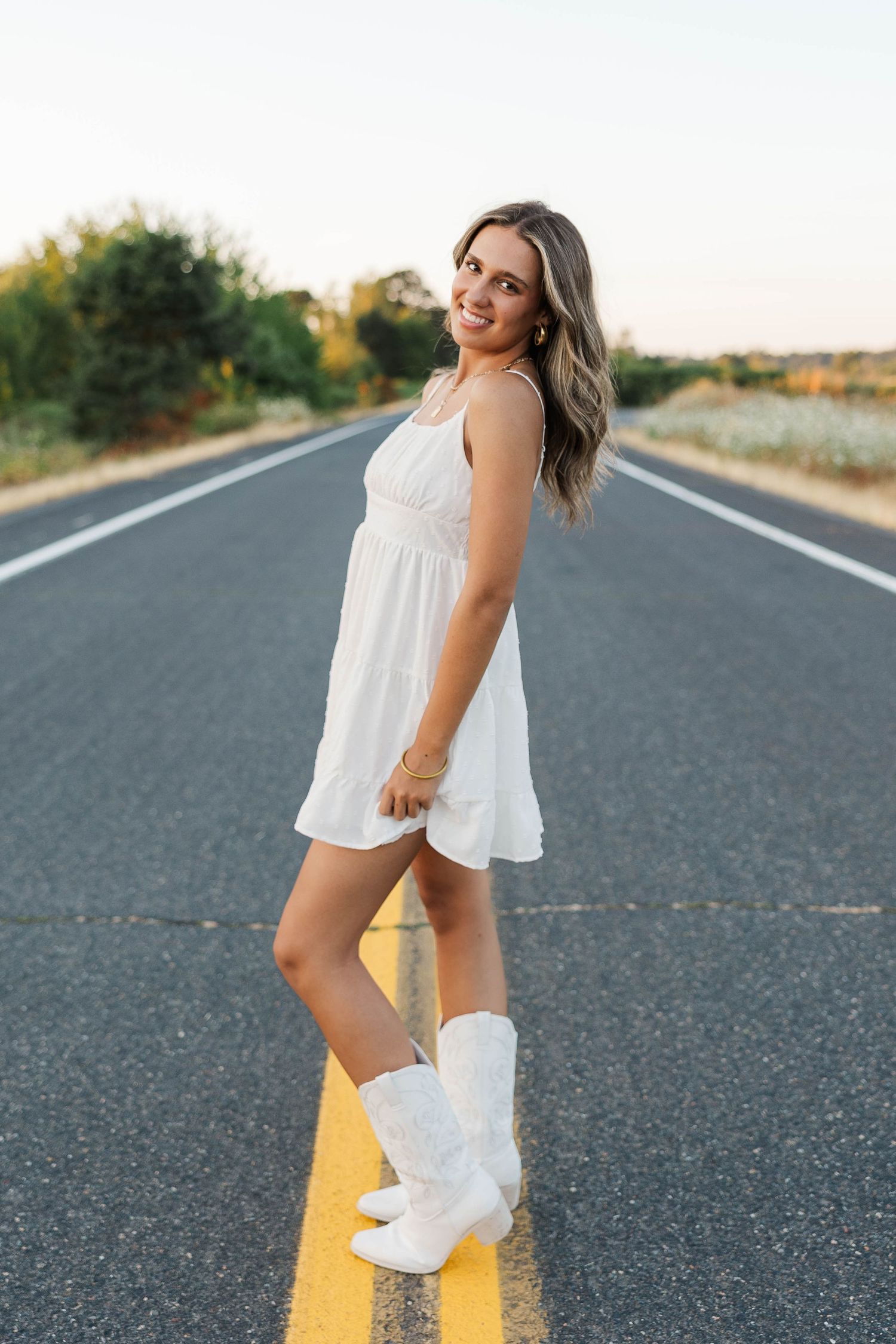 Young woman in white sundress and white cowboy boots standing on a road at sunset.