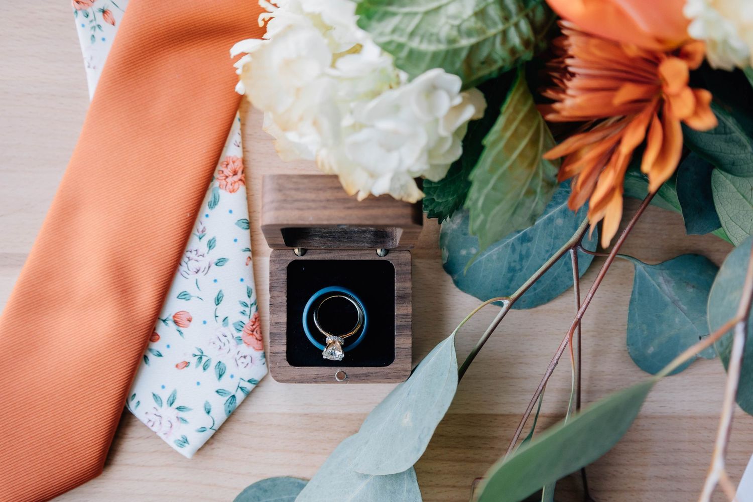 Wedding ring in wooden box beside orange tie, floral fabric, and orange and white flower bouquet on wood surface.