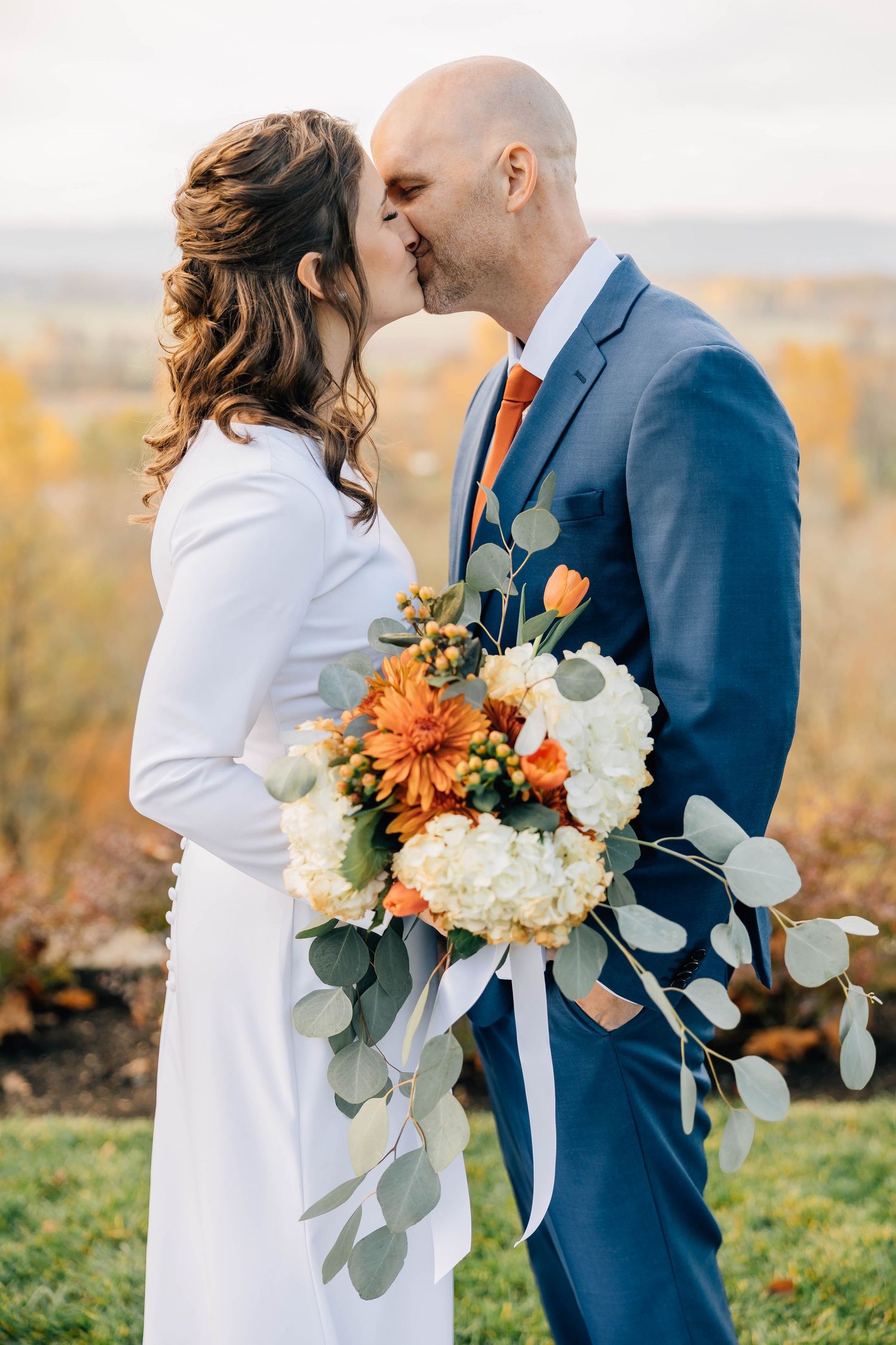 Bride and groom kissing outdoors in fall, holding orange and white bouquet, groom in navy suit with orange tie.