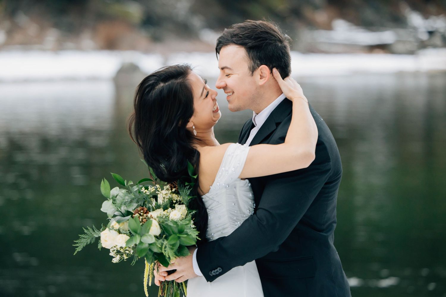 Bride and groom embracing and smiling by a lake, bride holding white and greenery bouquet, groom in black suit.