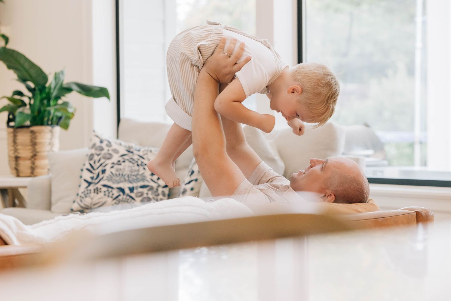 Mother lying on bed lifting happy toddler in the air in a bright sunlit living room.