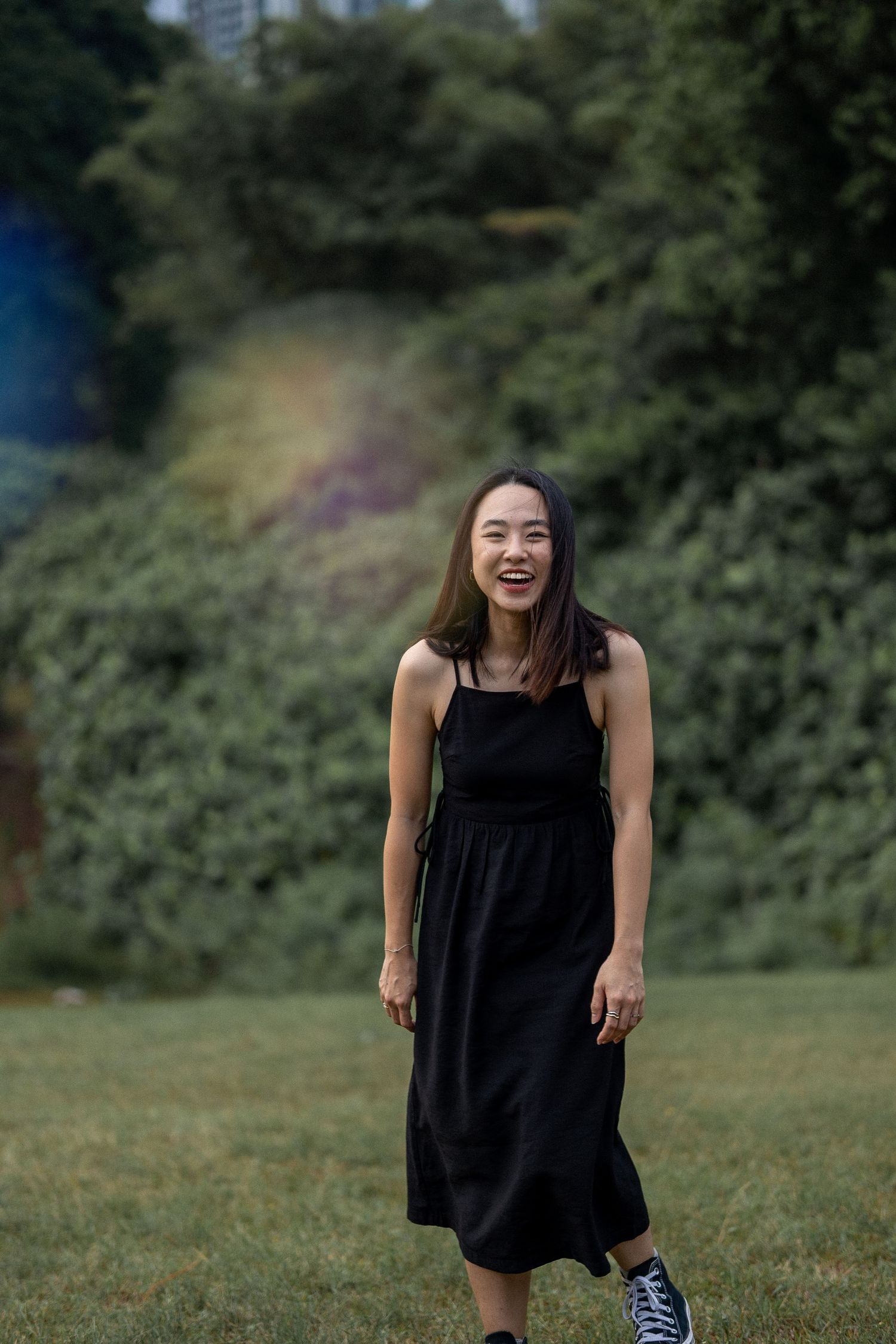 A person in a black sundress and sneakers stands joyfully on a grassy lawn with trees in the background.