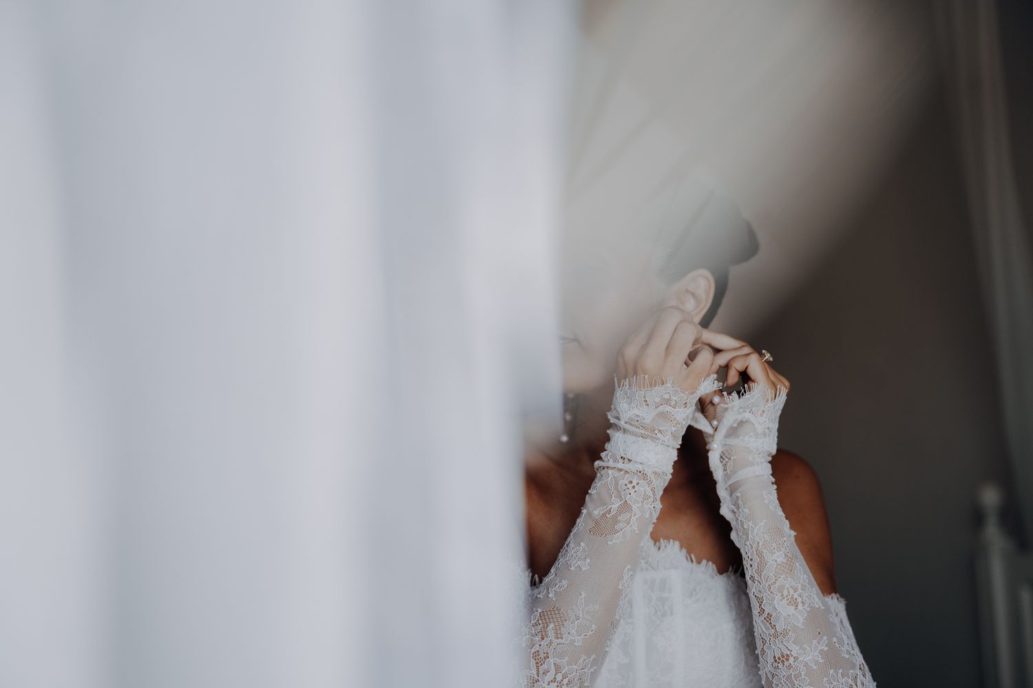 Bride in lace wedding dress and gloves adjusting veil near sheer white curtains in soft natural light.